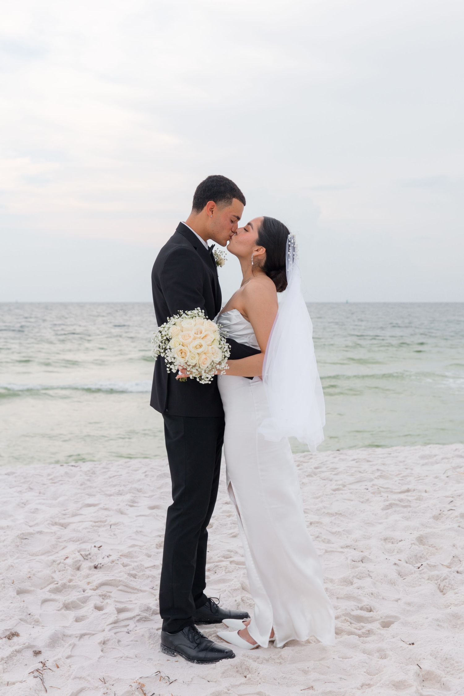 Newlyweds share a romantic kiss on a white sandy beach with ocean backdrop.
