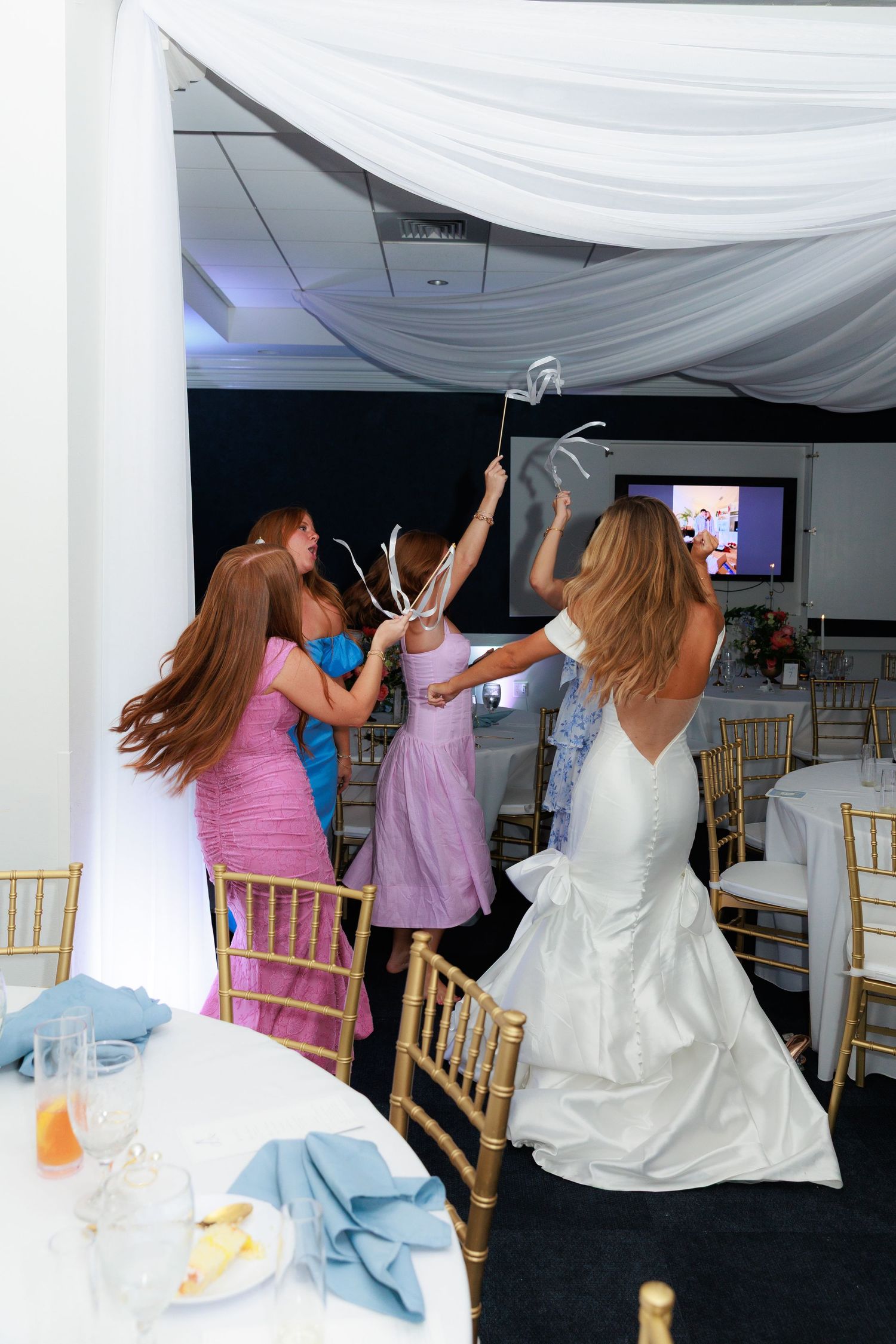 Group of people dancing and celebrating in formal attire at an indoor event with draped white fabric decor.
