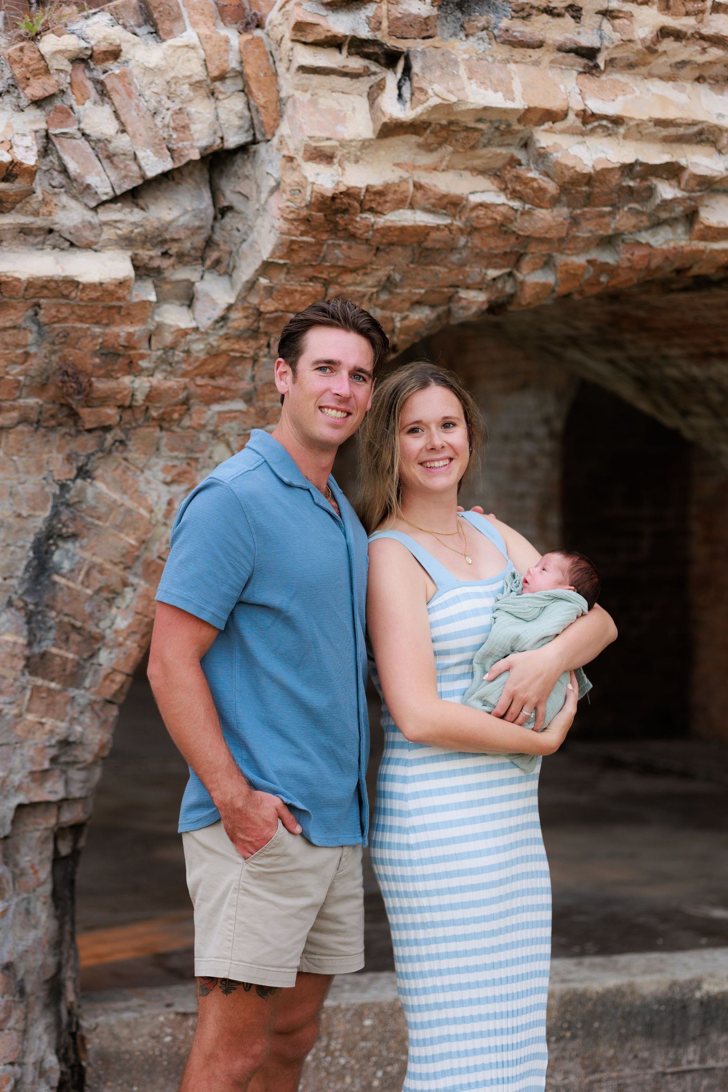 A couple stands with their baby in front of a rustic stone archway wearing coordinating blue and white outfits.