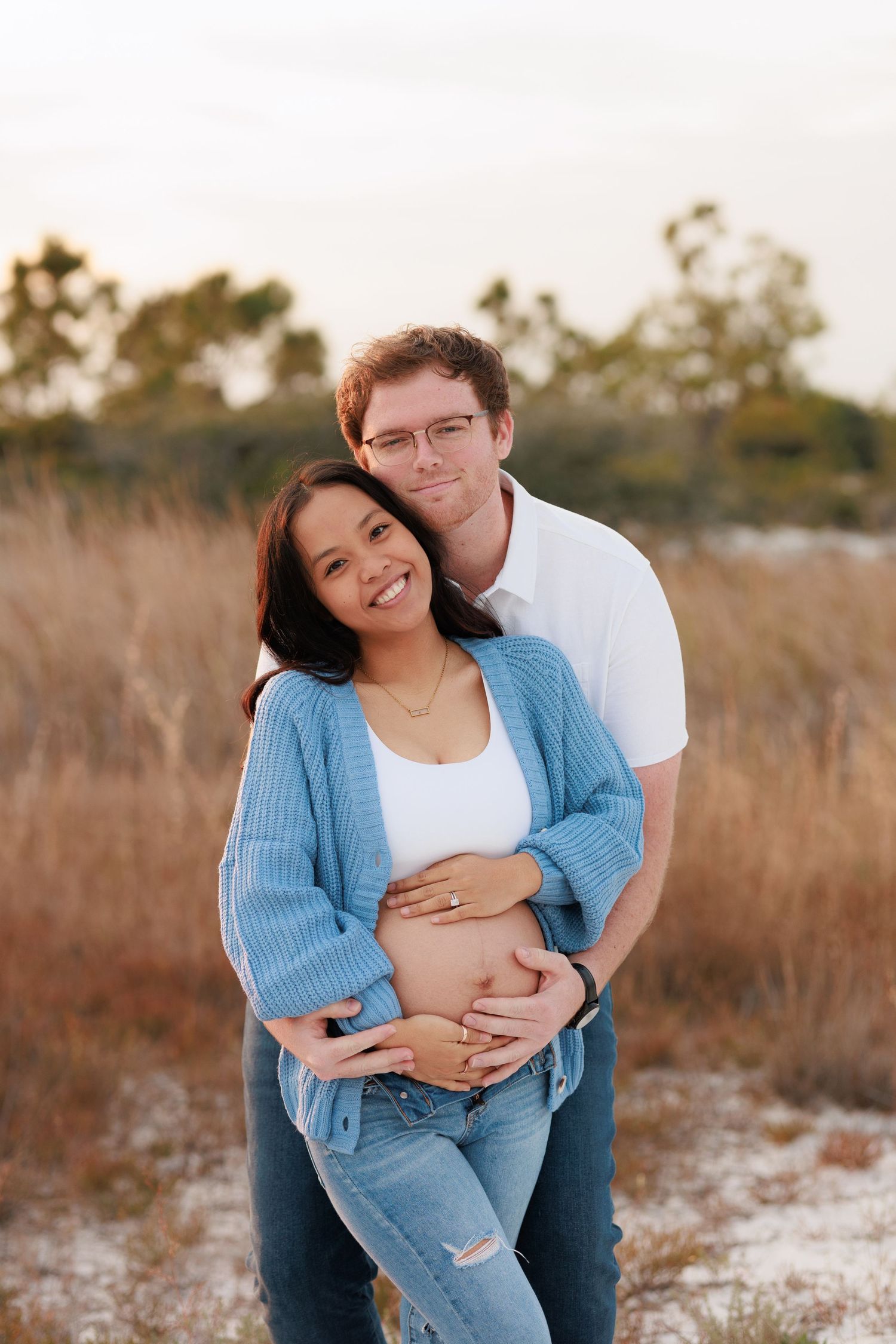 A couple embraces outdoors in casual attire during a sunset photoshoot in a natural setting.