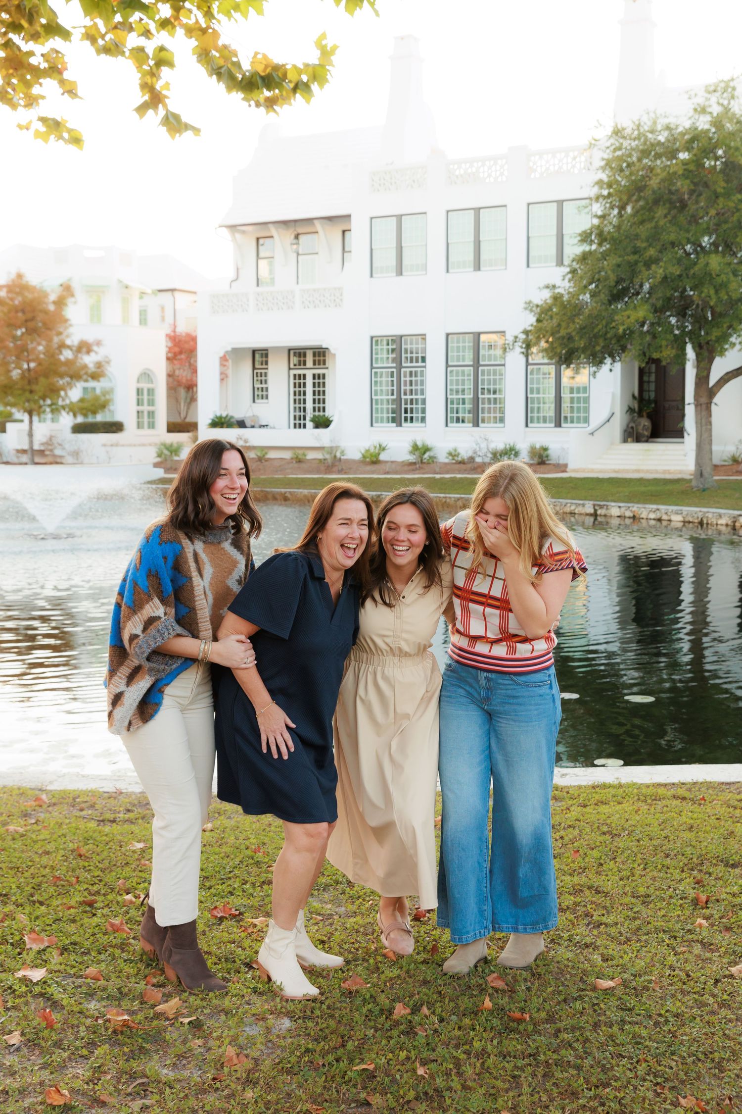 Four friends laugh together on grass near a pond and white building during golden hour.