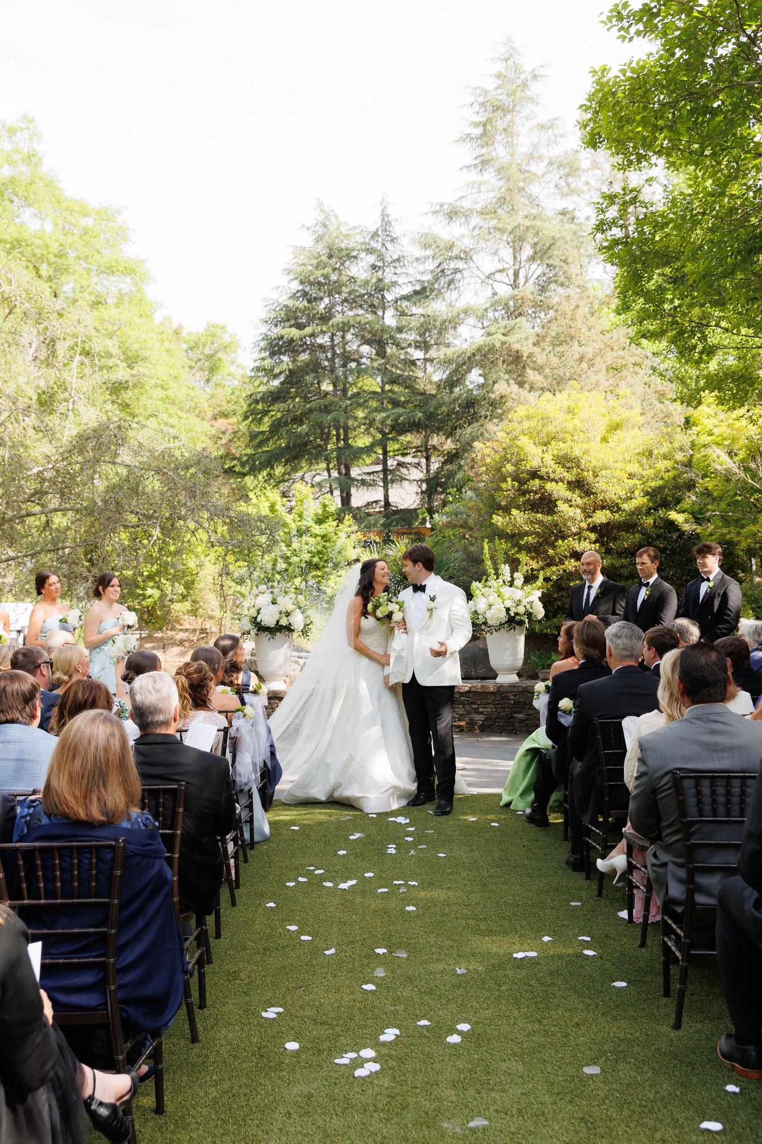 Bride and groom share a kiss at outdoor garden wedding ceremony with guests seated on black chairs along grass aisle.