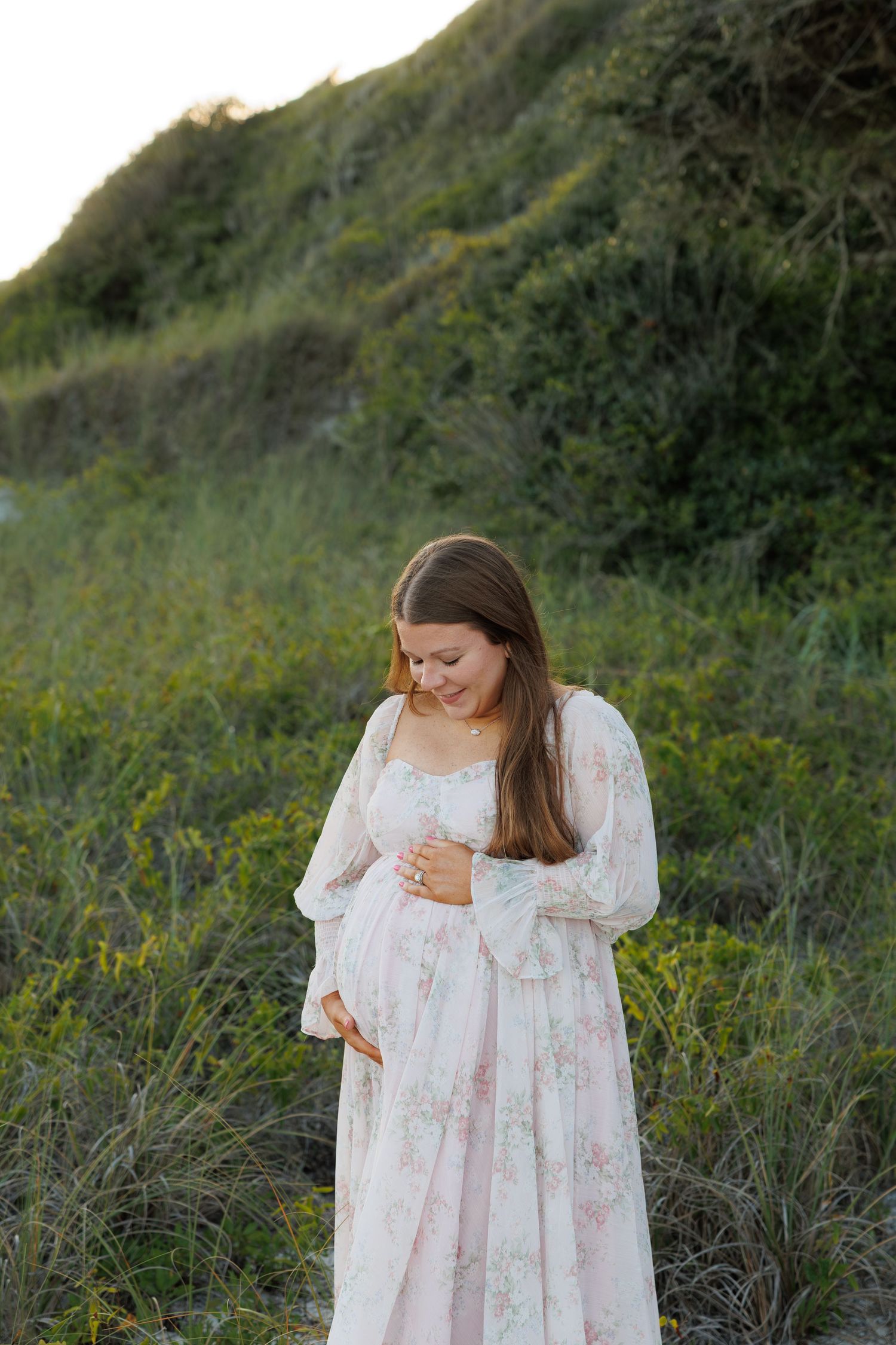 Pregnant woman in floral maxi dress holding baby bump outdoors near green hillside.