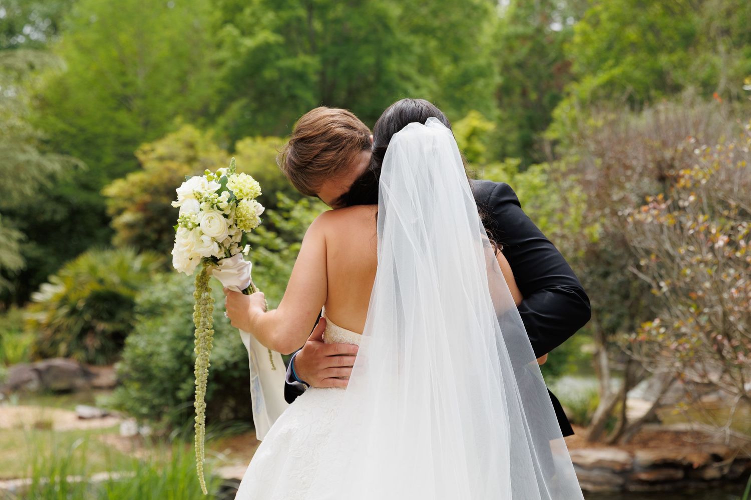 Bride and groom embracing from behind in a garden, bride holding white bouquet and wearing veil.