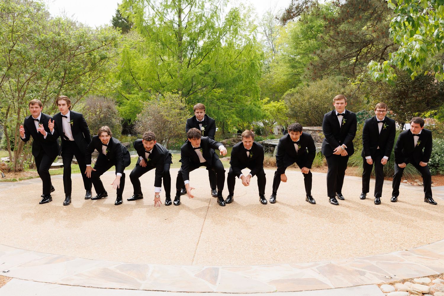 Groomsmen in black tuxedos strike fun crouching poses on a stone patio surrounded by trees at a wedding.