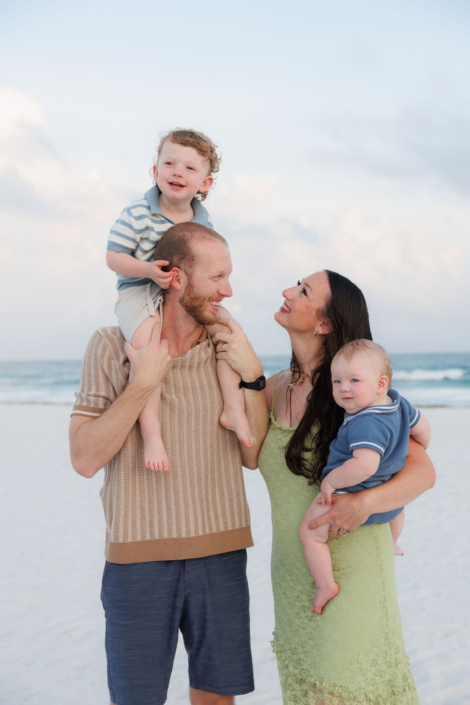 A happy family of four poses on a beach, with dad holding toddler on shoulders and mom cradling baby.
