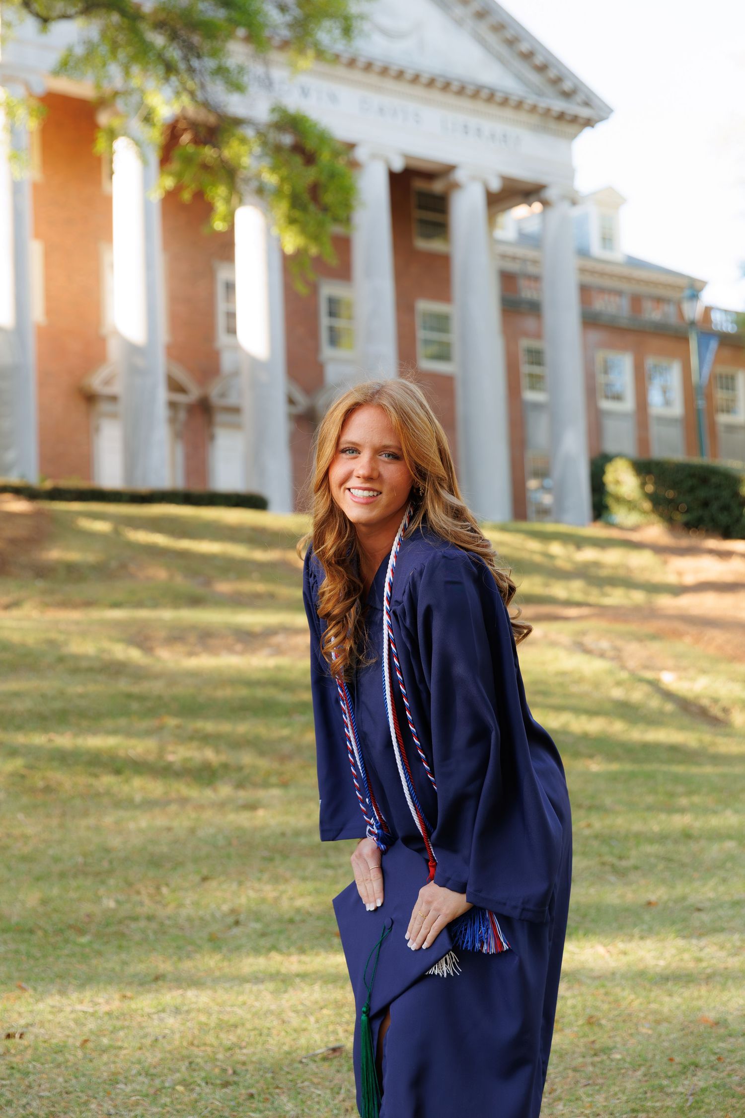 Happy female graduate in navy cap and gown poses on campus lawn in golden hour sunlight.