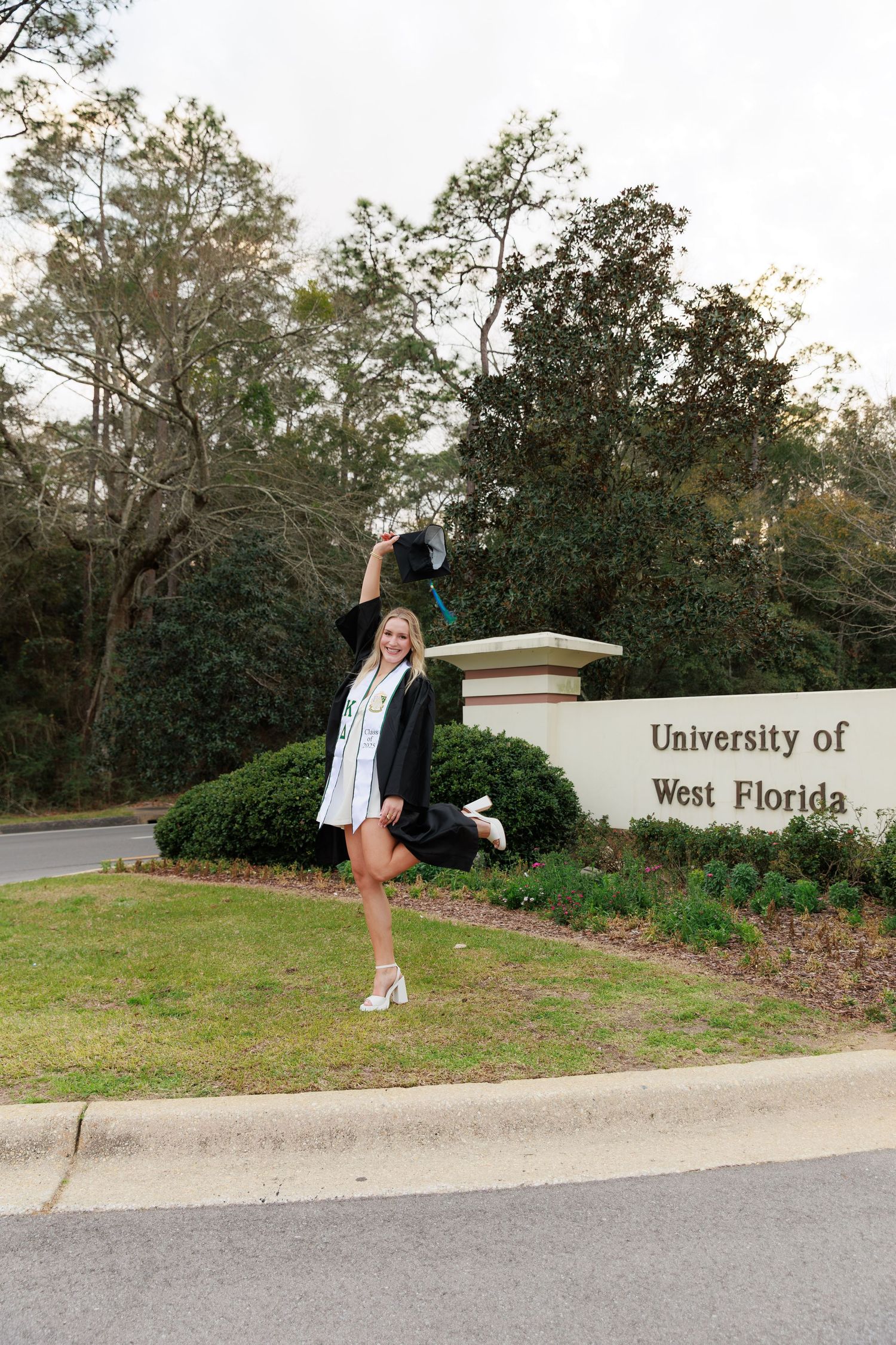 Smiling female graduate in cap and gown celebrating in front of University of West Florida entrance sign.