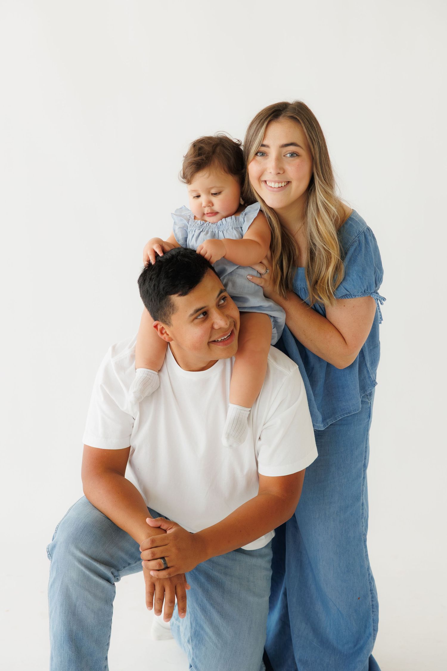 Family portrait with father seated, mother in blue jumpsuit and toddler on his shoulders, white background.