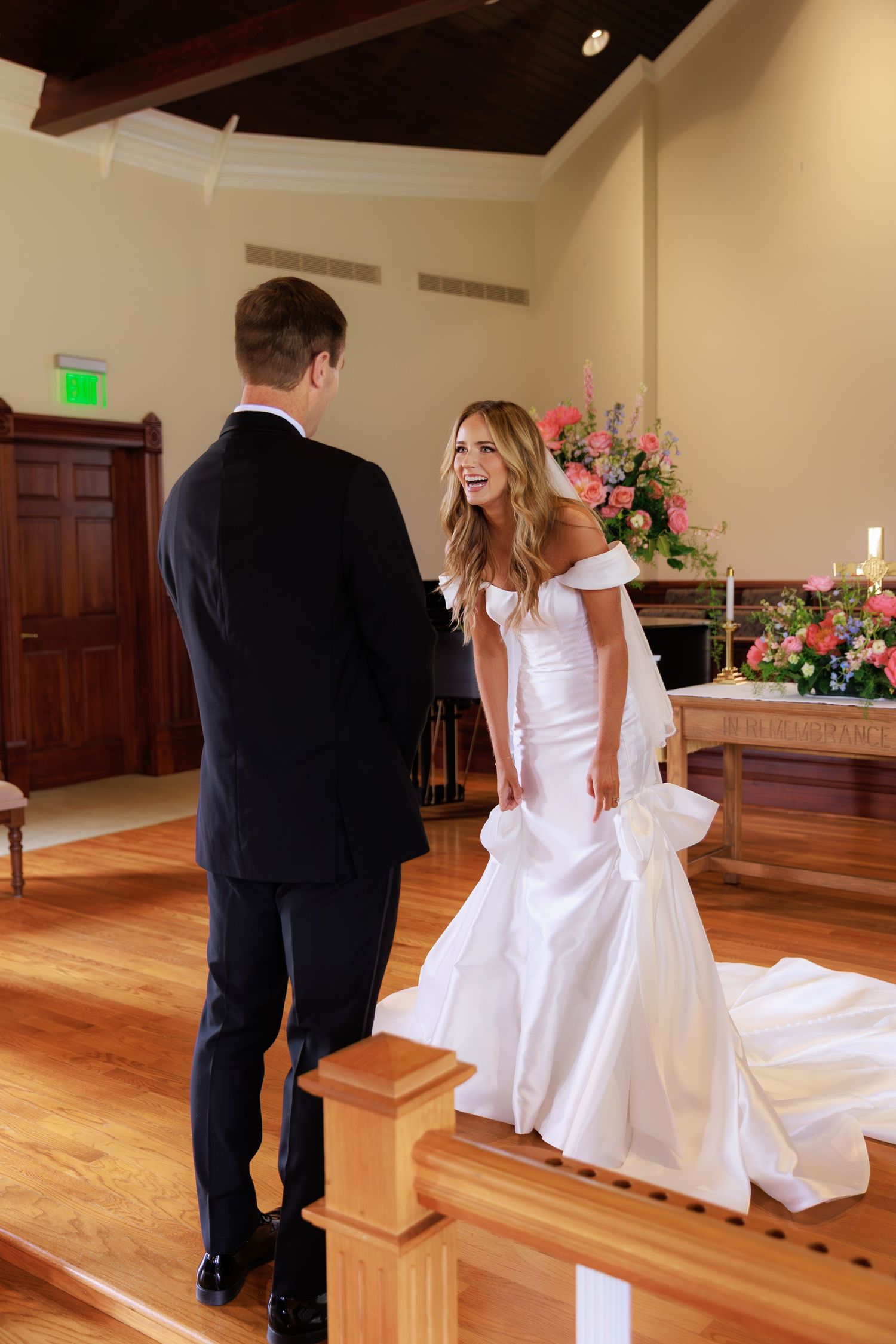 A couple stands facing each other during their wedding ceremony in a church with wooden floors and floral decorations.