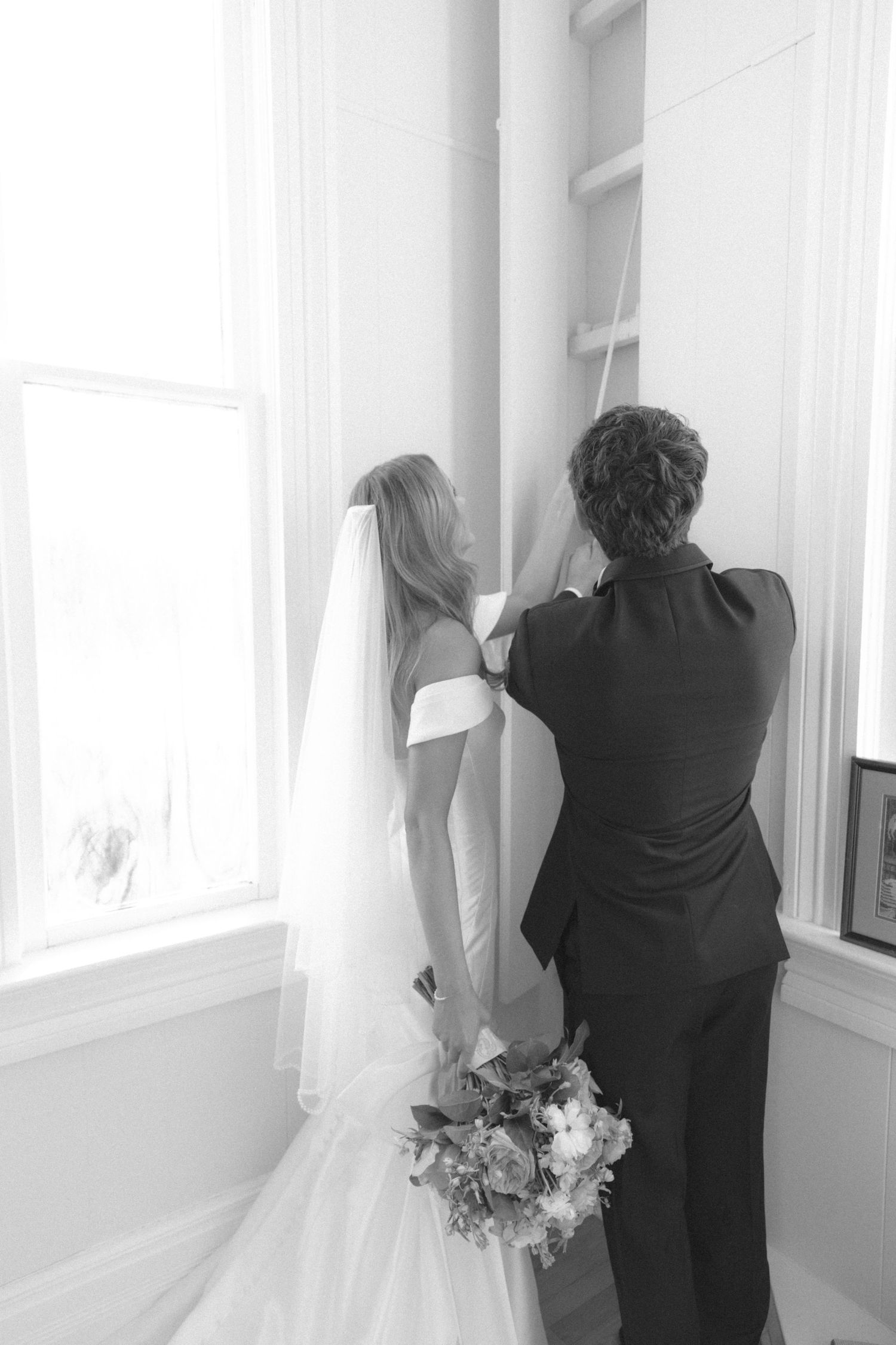 Black and white photo of a bride in wedding dress holding bouquet looking out window with companion.