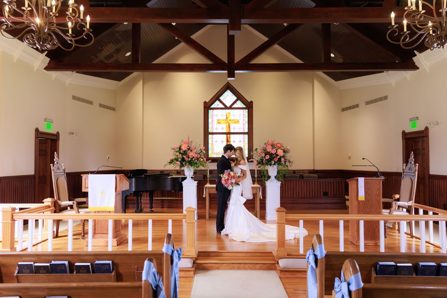 A traditional chapel interior with wooden pews, chandeliers, and pink floral arrangements at the altar.