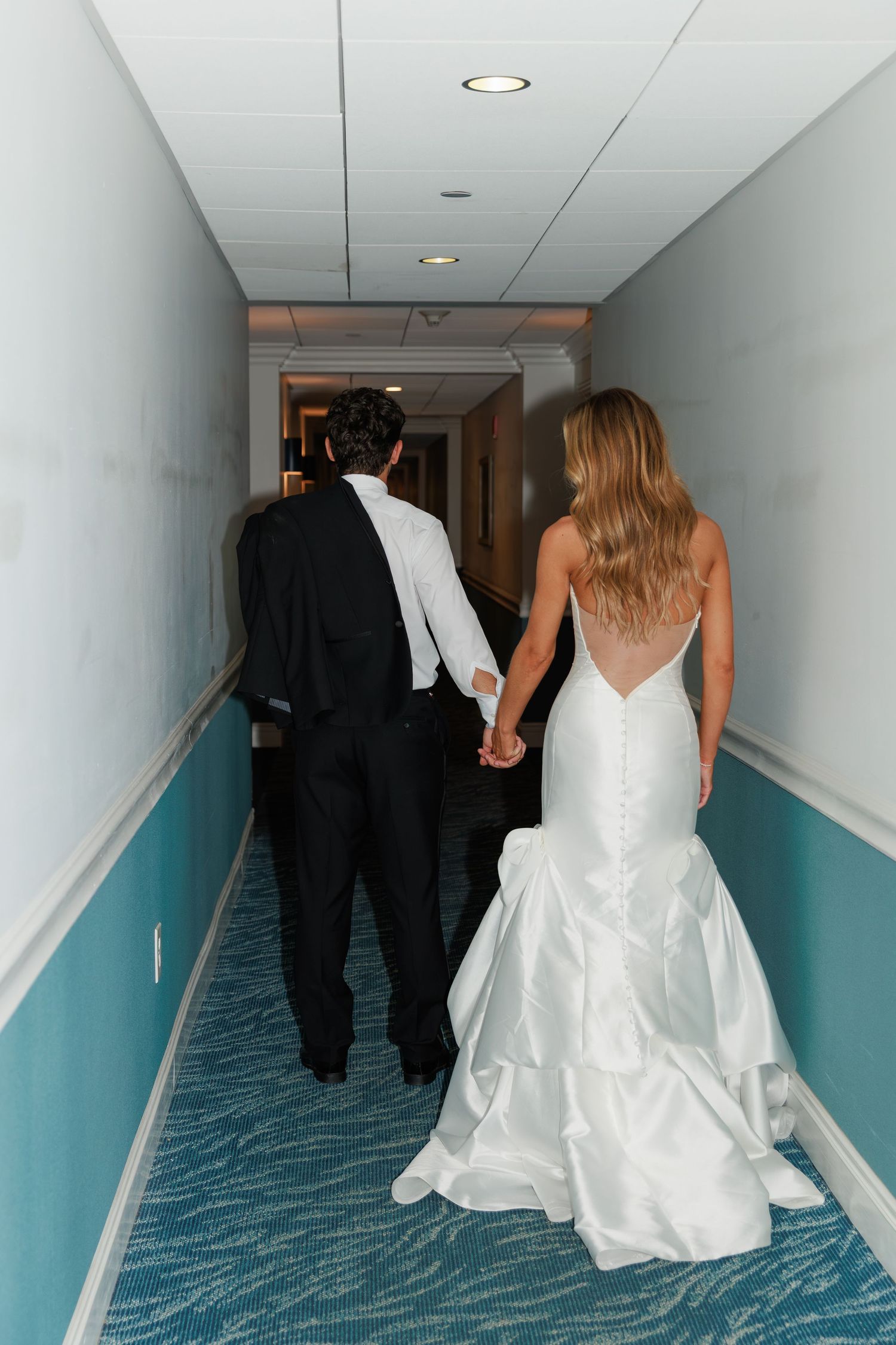 Wedding couple walking down hotel hallway with turquoise walls and carpet.