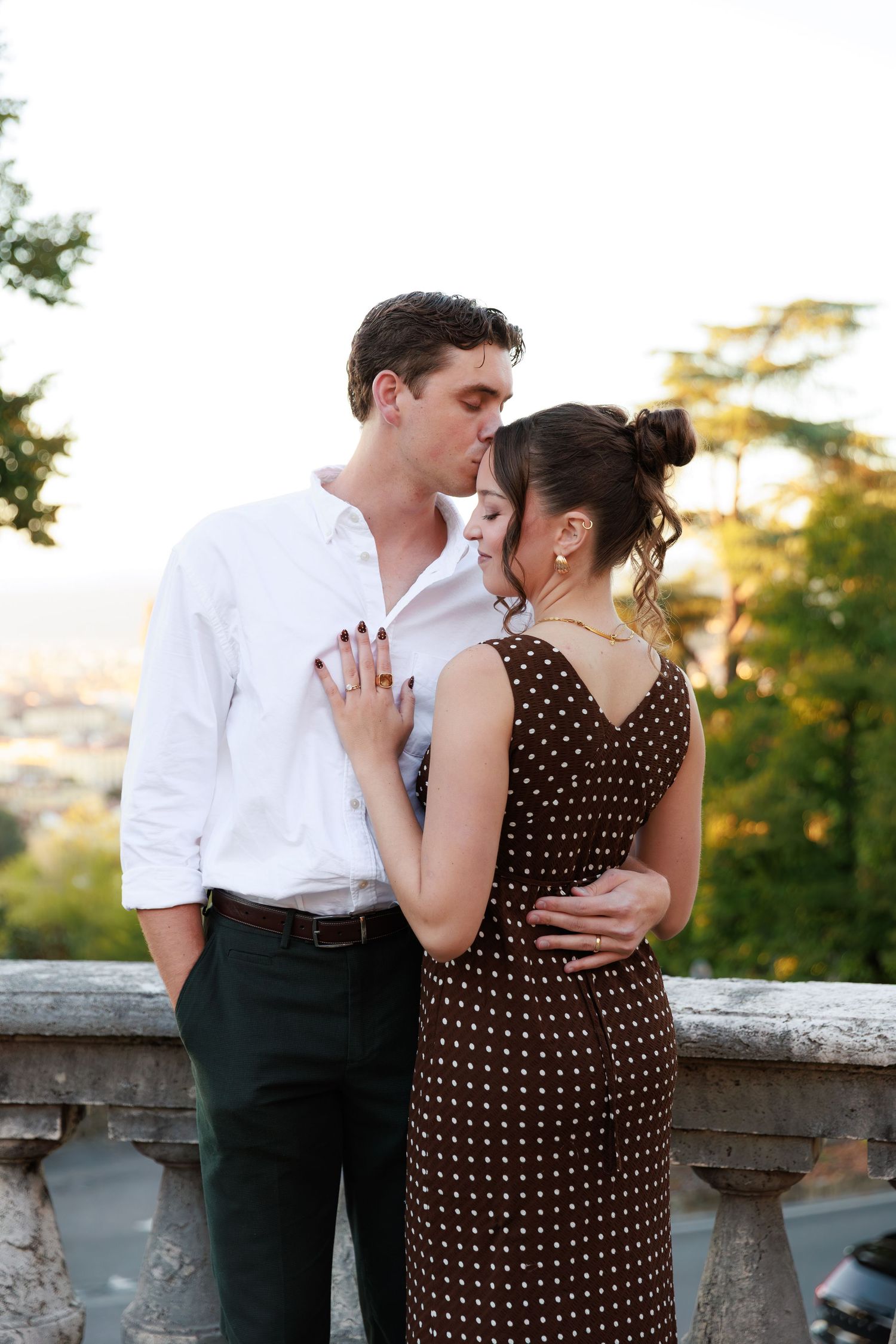 Romantic couple embracing on a scenic balcony, man in white shirt kissing woman in brown polka dot dress at sunset.