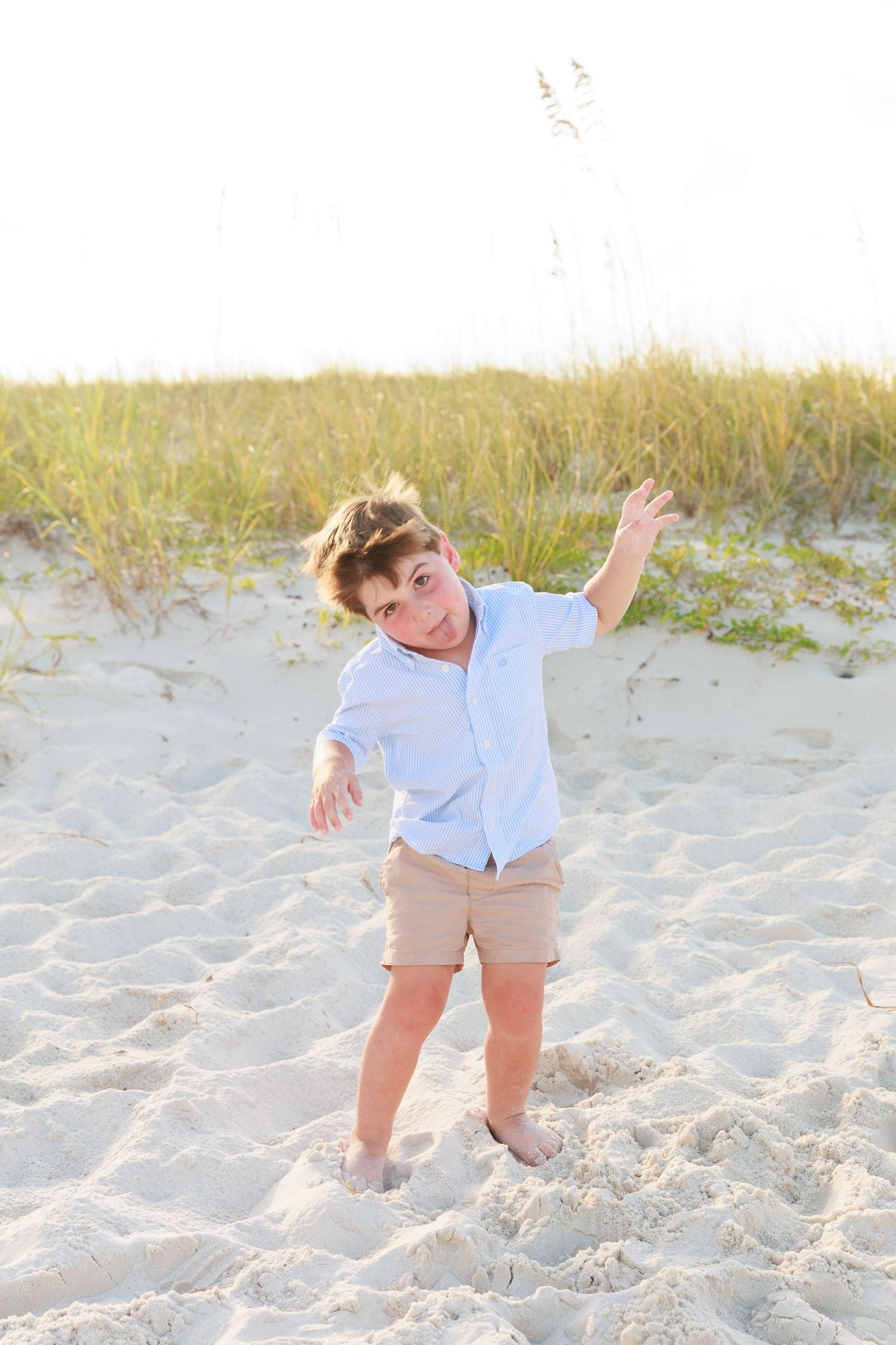A young boy in a light blue shirt and khaki shorts plays on a sandy beach with sea oats in the background.