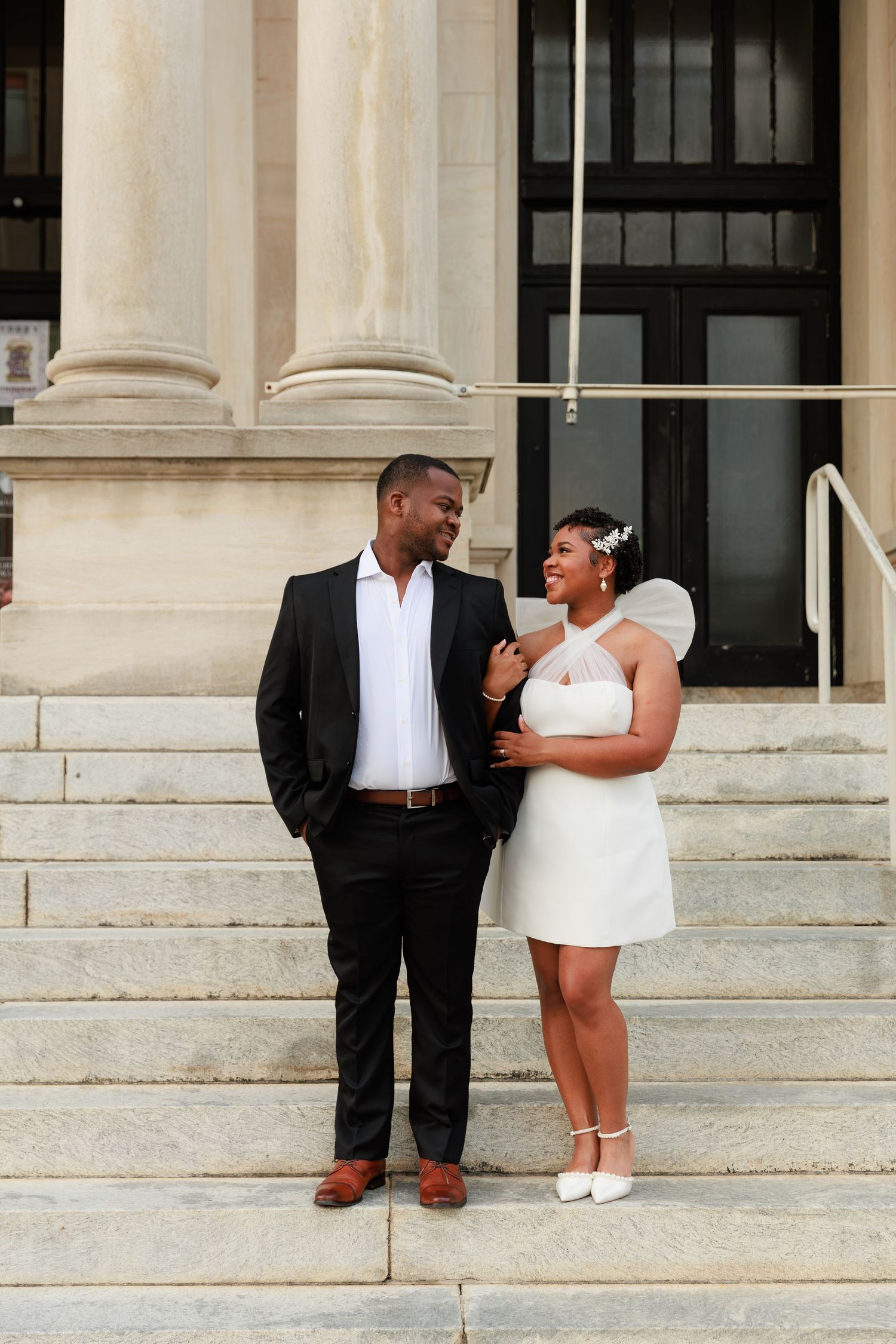 Couple in wedding attire smiling at each other on courthouse steps, bride in white mini dress, groom in black suit.