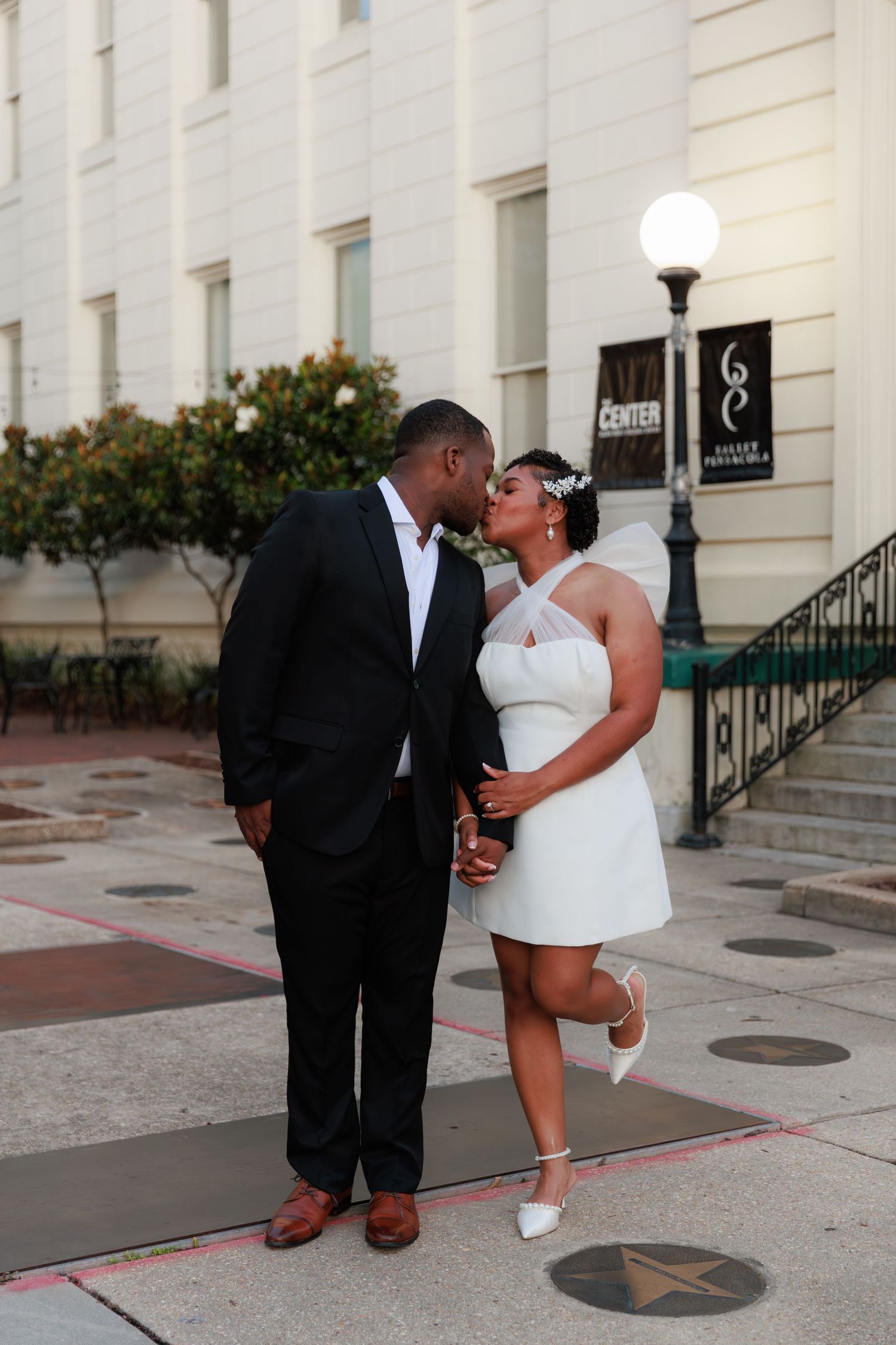 A bride and groom share a kiss outside a classical building, she in a short white halter dress with veil, he in a black suit.