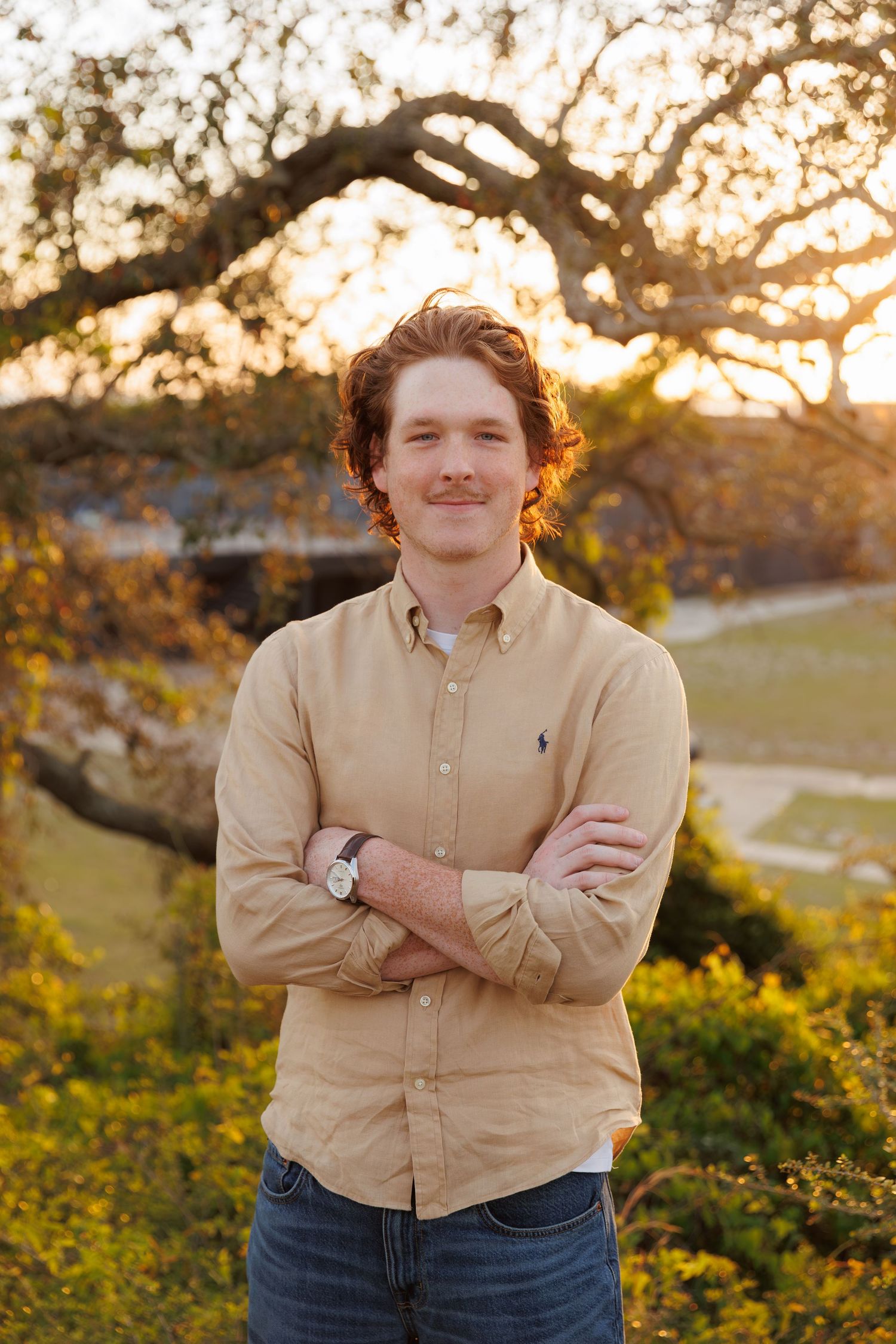 Young man with crossed arms smiling outdoors at golden hour, wearing a tan Ralph Lauren shirt and jeans.