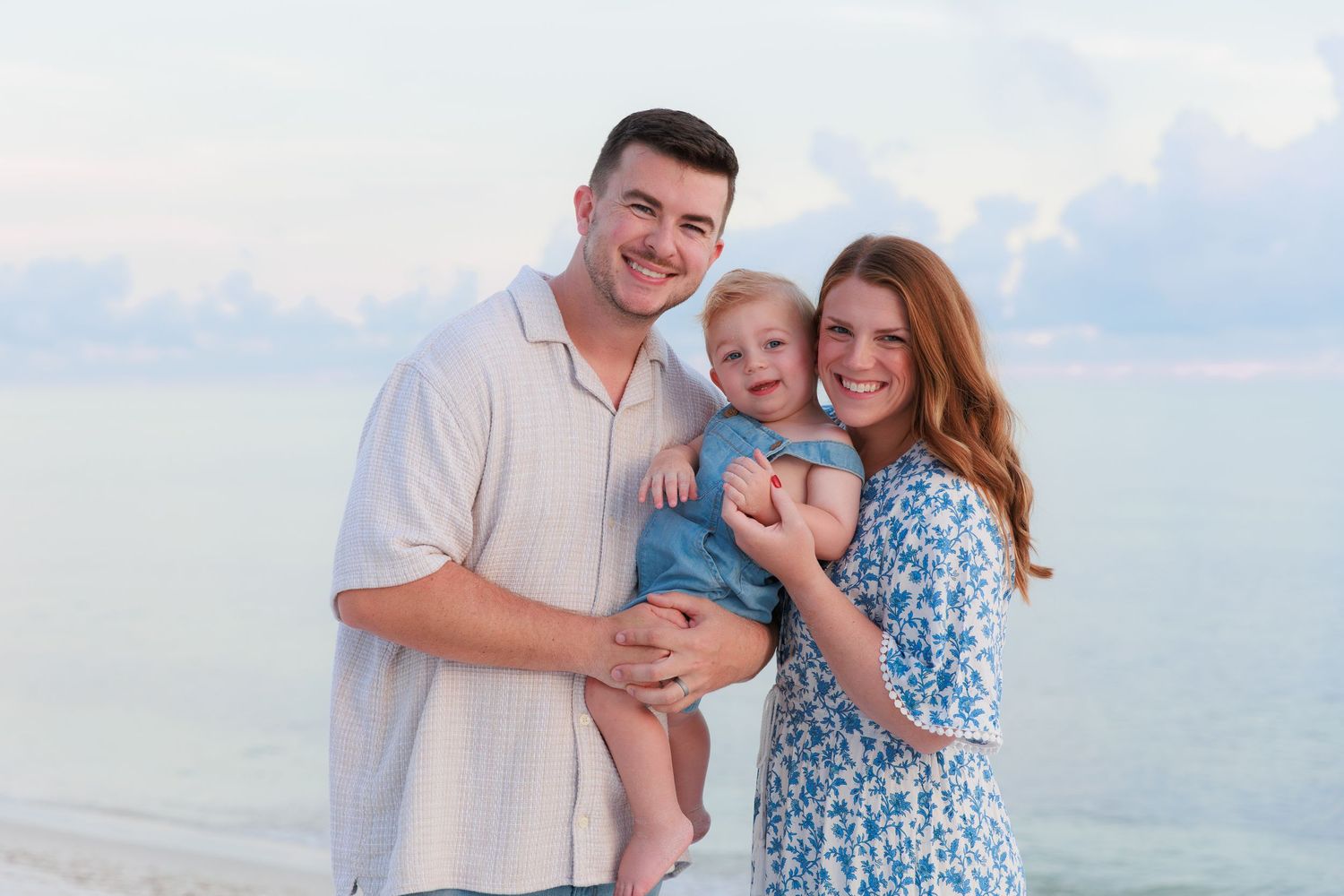Happy family of three posing on a beach, parents holding toddler with calm ocean waters in the background.