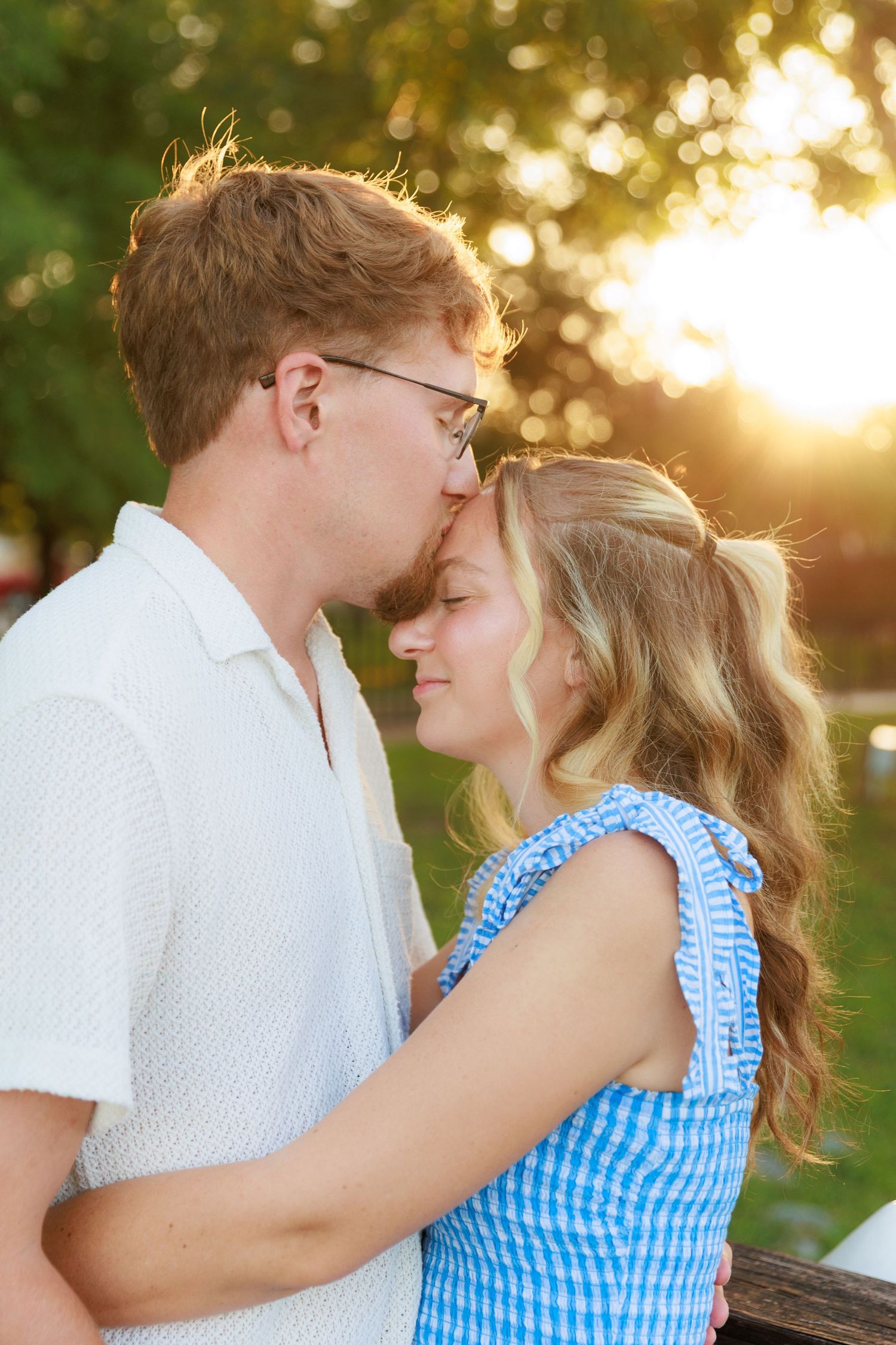 Man kissing woman on forehead at golden hour outdoors, both embracing romantically in a park.