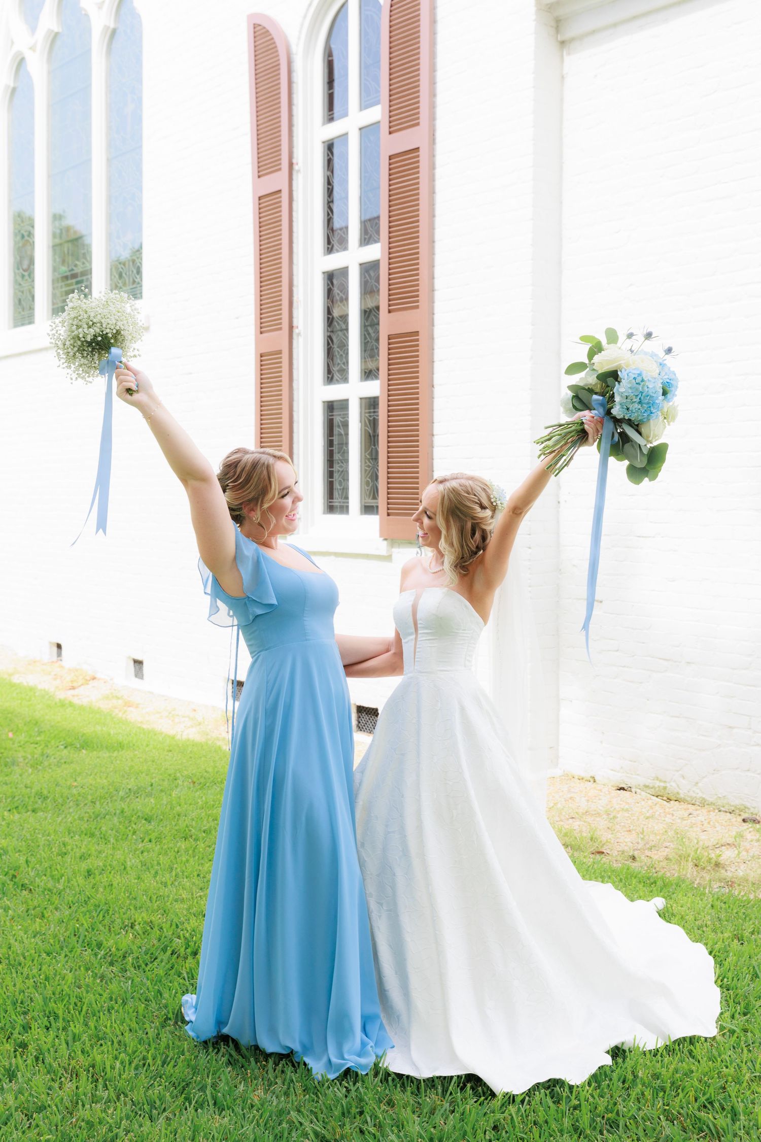 Bride and bridesmaid raise bouquets triumphantly outside white church building.