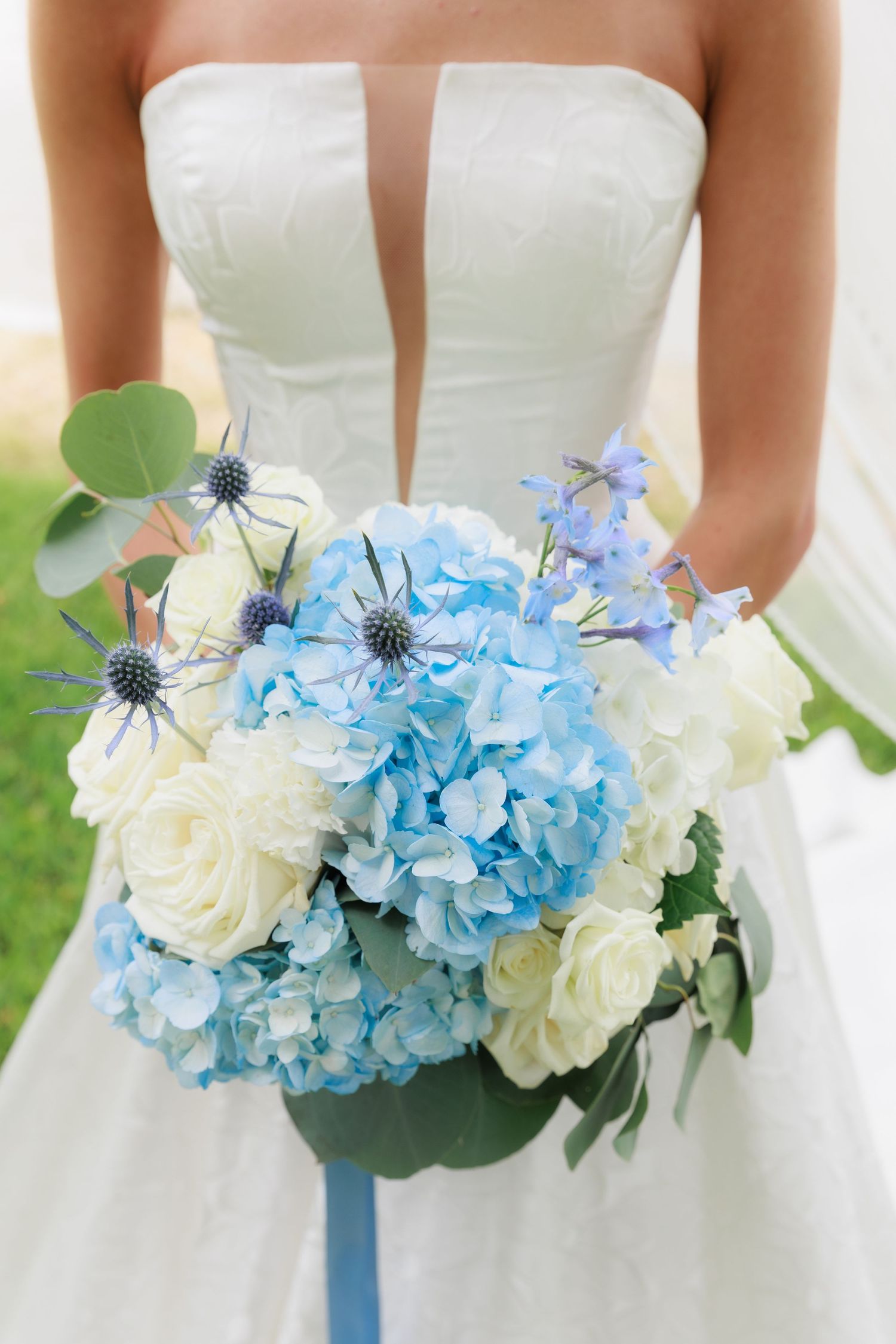 A wedding bouquet featuring blue hydrangeas and white roses against a white dress.