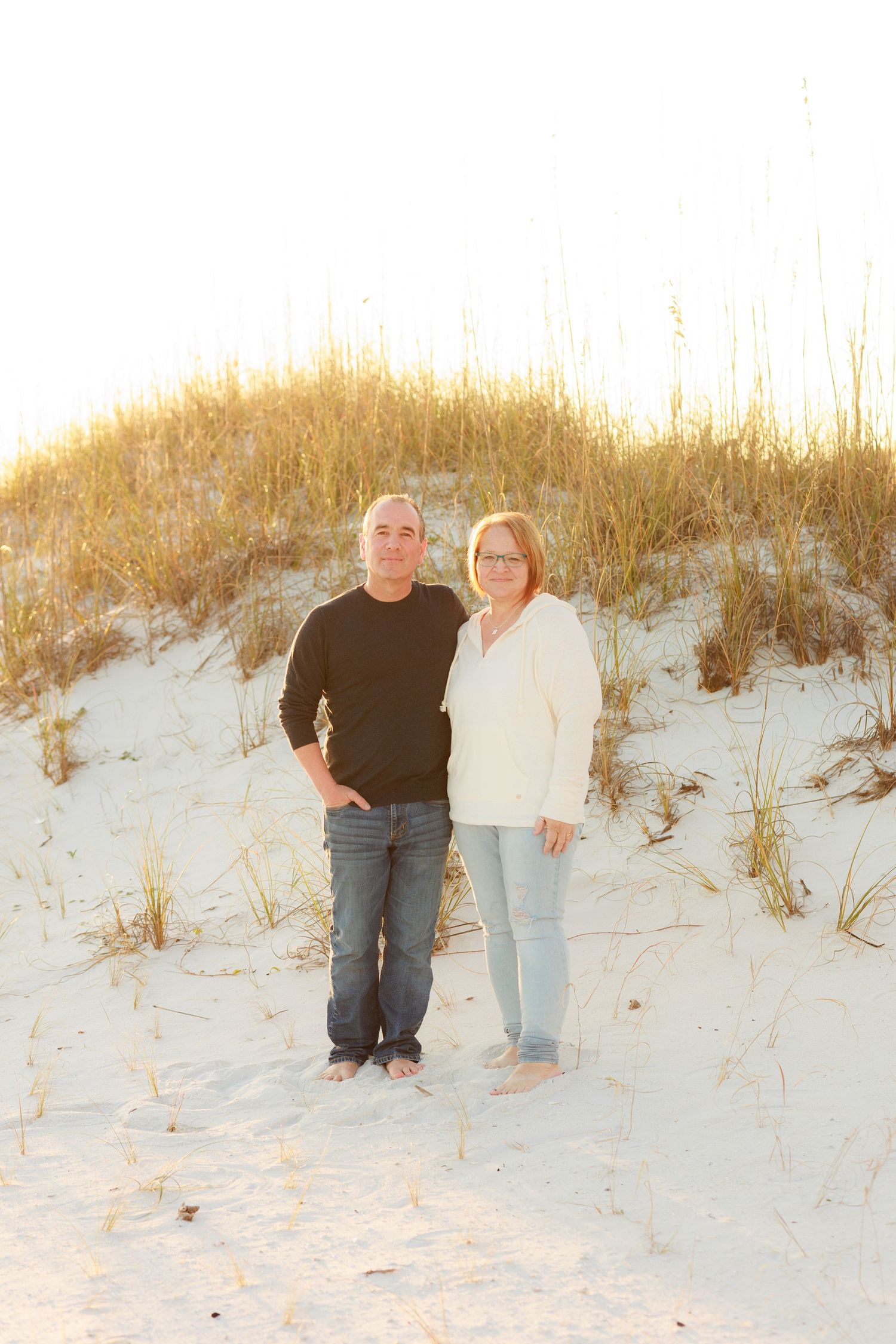 A couple stands together on a white sandy beach dune during golden hour sunset.