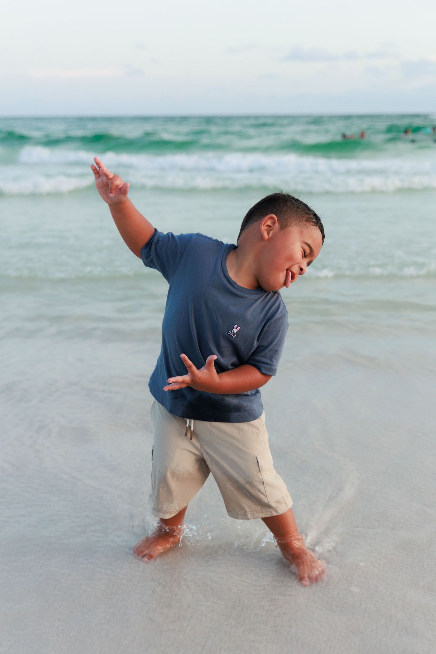 A young boy in a blue shirt plays joyfully in the shallow ocean waves at the beach.