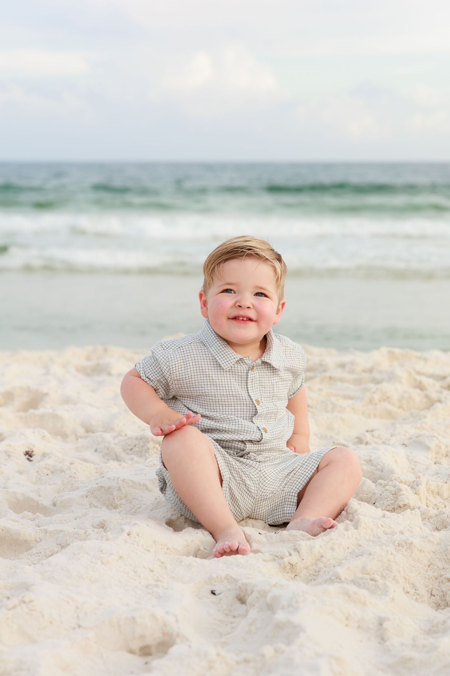 A small child wearing a checkered romper sits happily on a white sandy beach with ocean waves in the background.