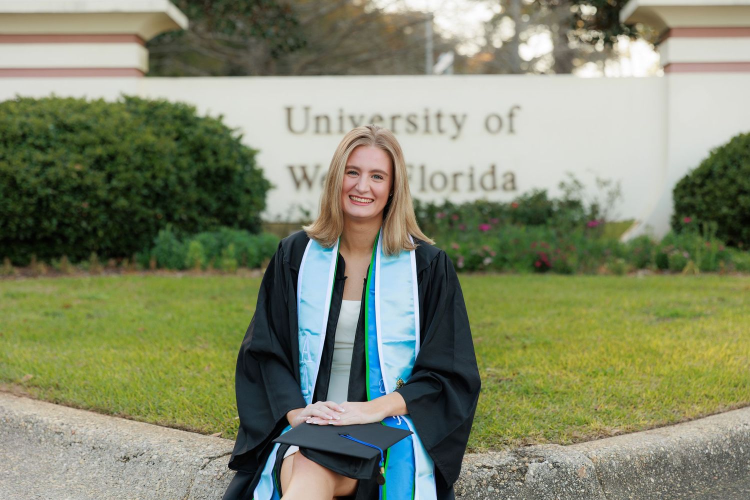 Graduate in black cap and gown with teal stole stands in front of University of Florida sign during graduation photos.
