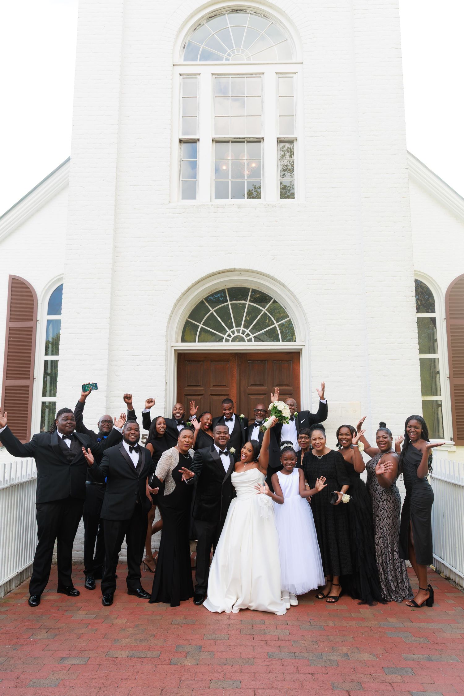 Wedding party celebrates outside a white church with arched windows and red brick patio during a joyous reception.