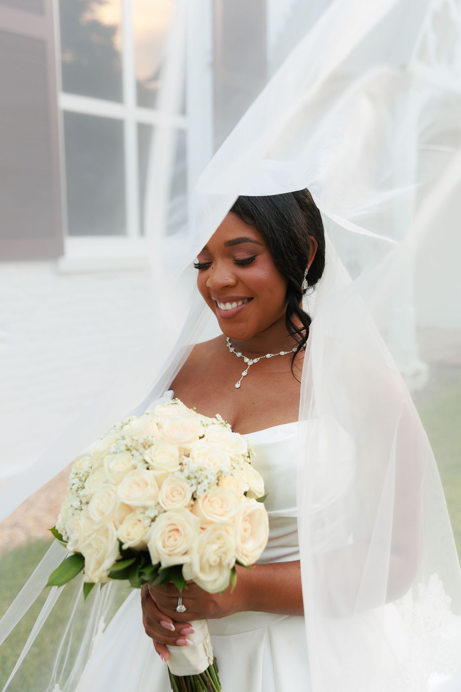 A radiant bride holds a bouquet of white roses while wearing an elegant veil and necklace.
