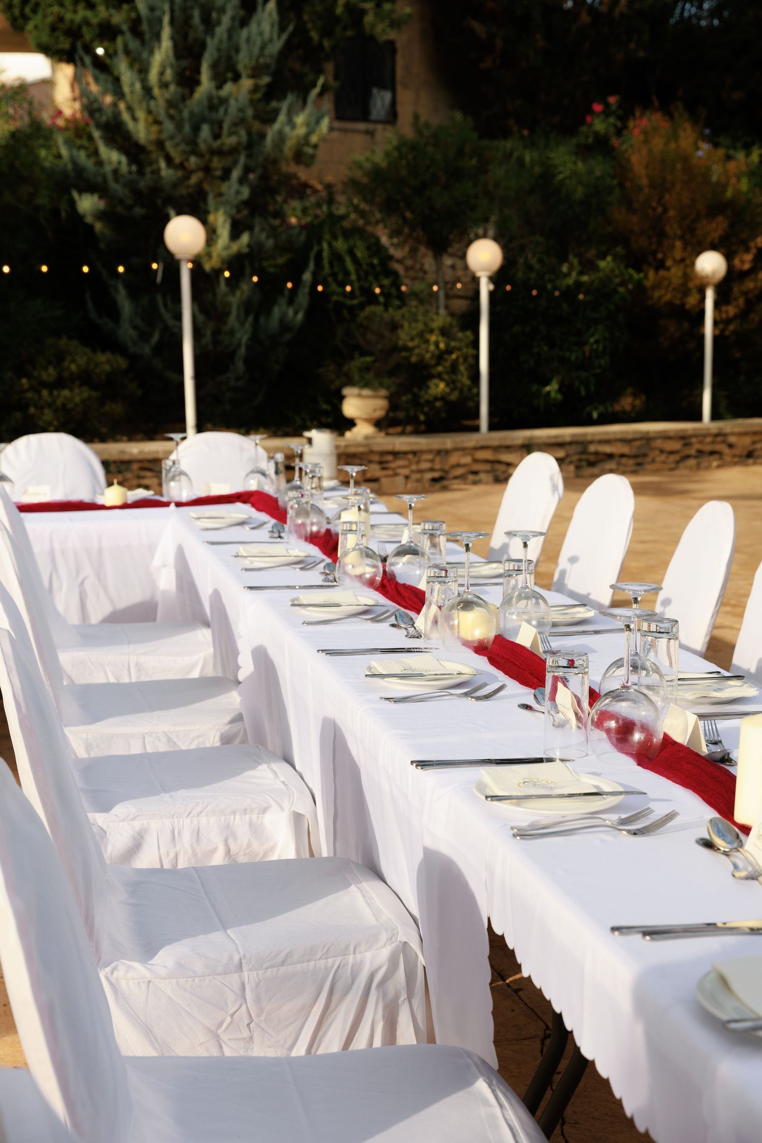 Long white dining tables set outdoors with elegant place settings and burgundy napkins for an elegant garden party.