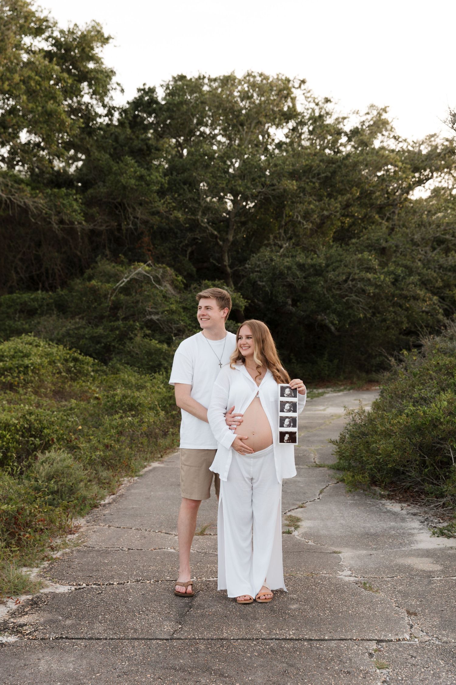 A couple in white outfits walking along a sandy beach path surrounded by green foliage.