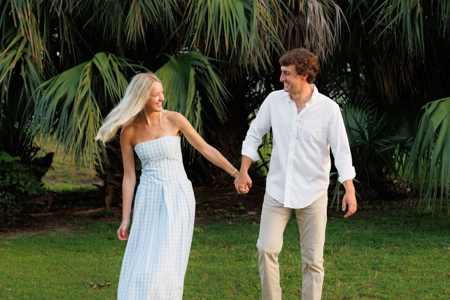 A smiling couple holds hands while walking on lush green grass with tropical palm trees in the background.