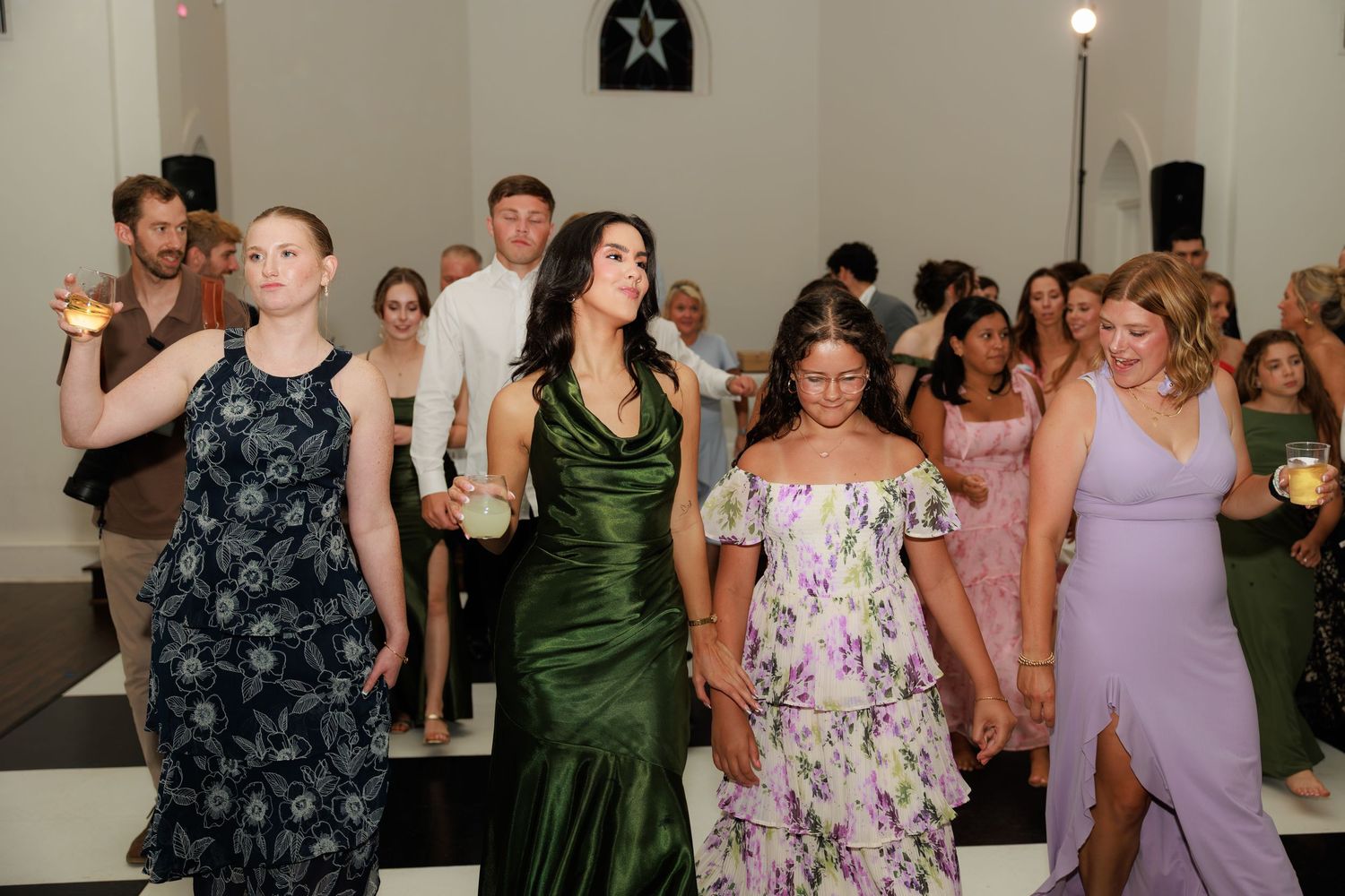 Women in elegant formal dresses dancing together at a wedding reception on a black and white checkered floor.