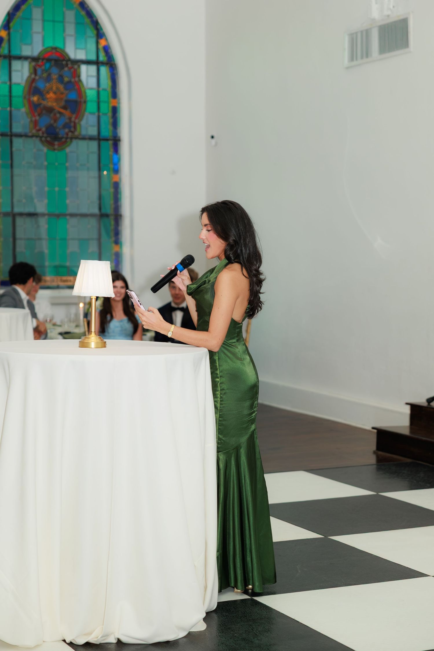 Woman in green satin gown delivers a speech at a wedding reception podium with stained glass windows in background.