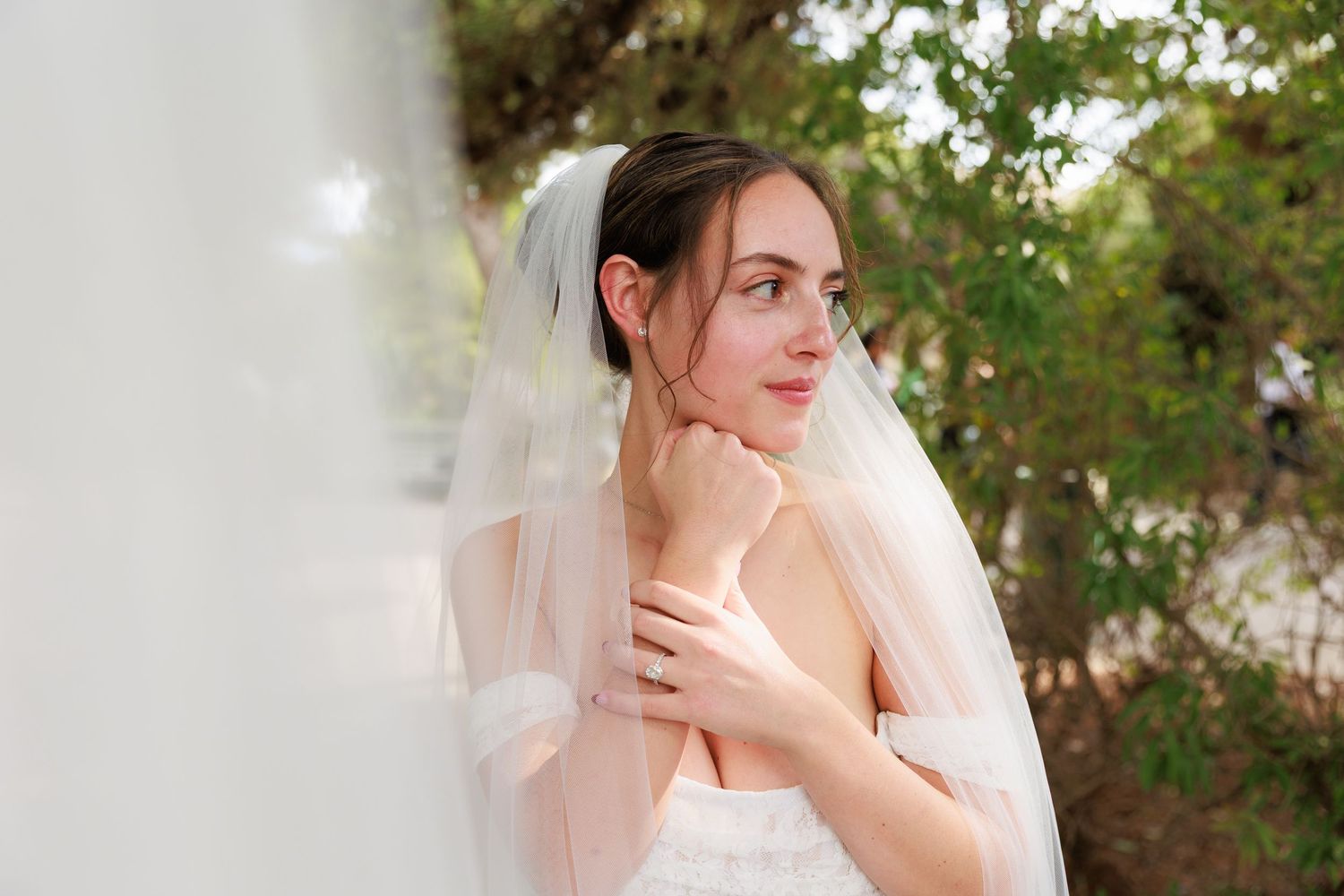 Bride in white veil poses thoughtfully against green foliage background in romantic wedding portrait.