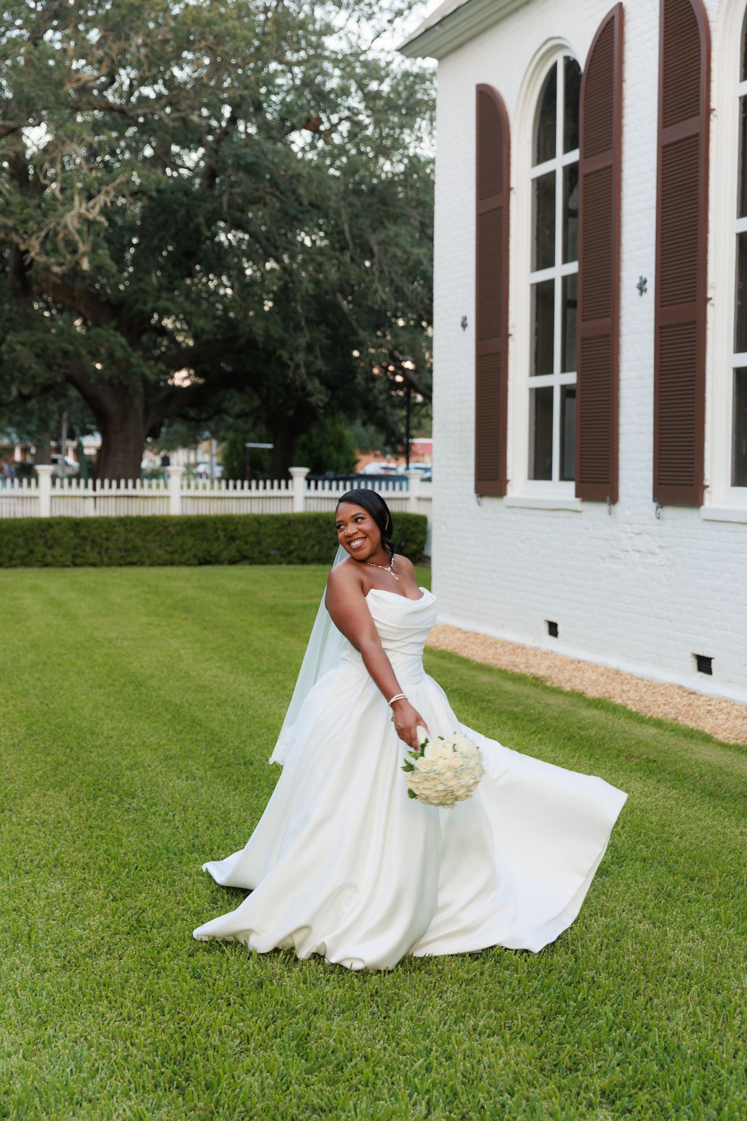 Bride in elegant white gown poses gracefully on manicured lawn with classic white building behind.