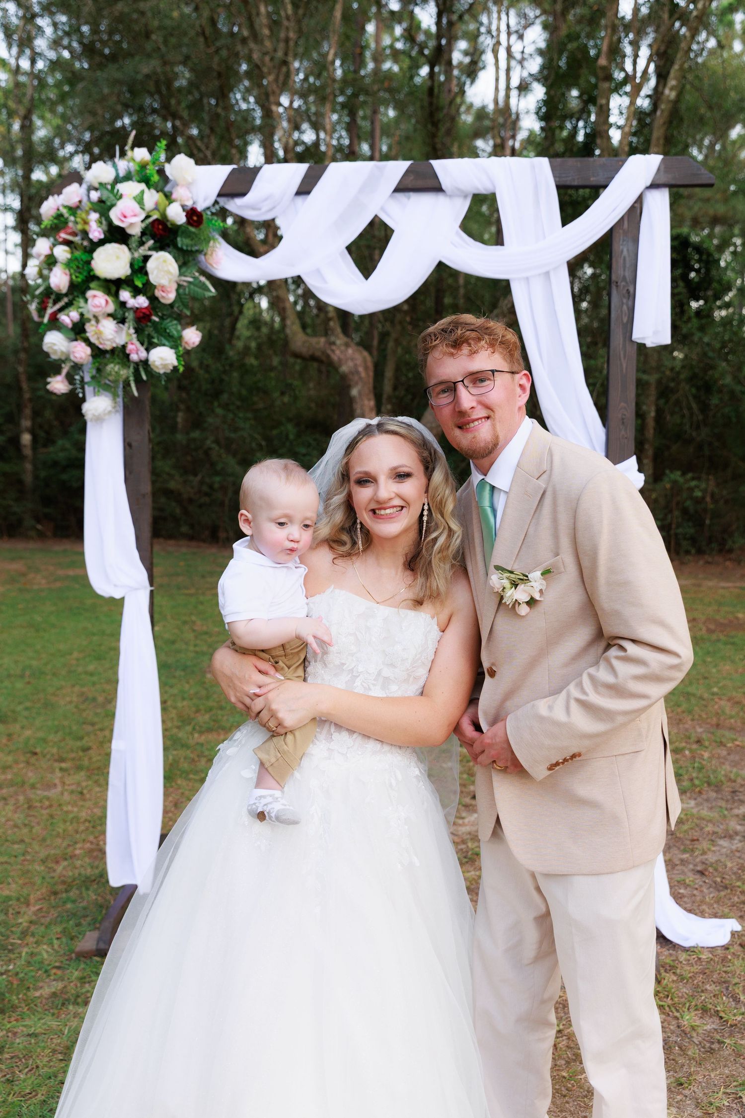 Wedding couple poses under white draped arch decorated with pink roses and greenery in outdoor garden setting.