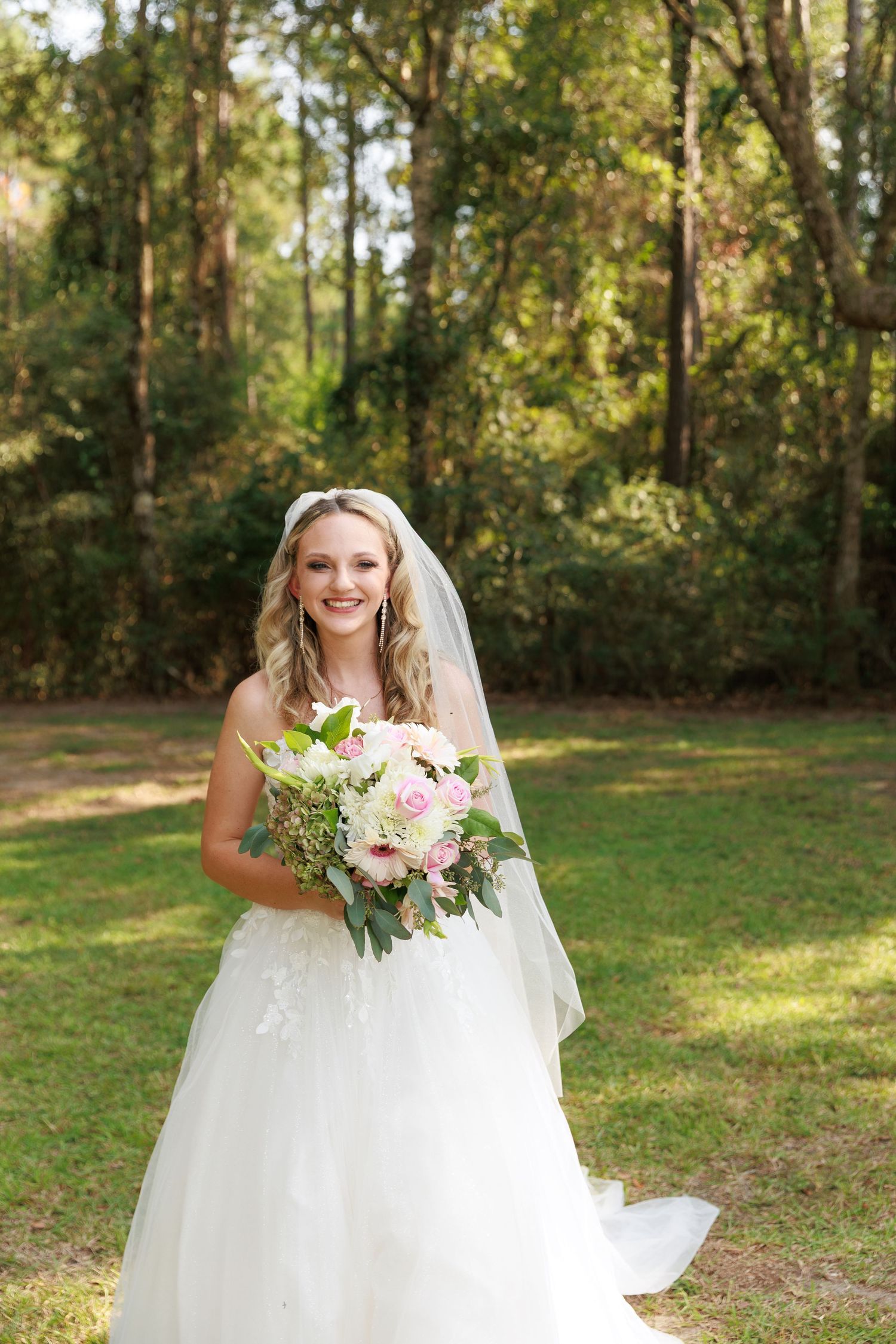 A bride in a flowing white wedding dress holds a pink and white floral bouquet in a sunlit outdoor forest setting.