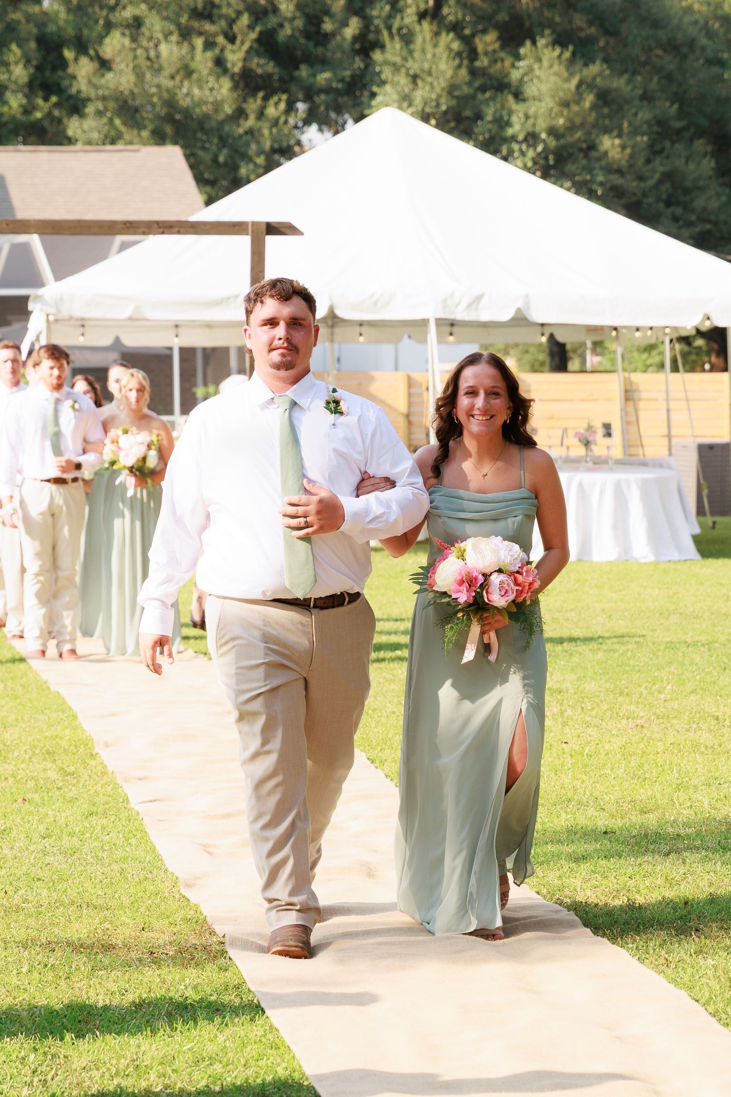 Wedding party members walk and laugh together on a sunny lawn with white tents in the background.