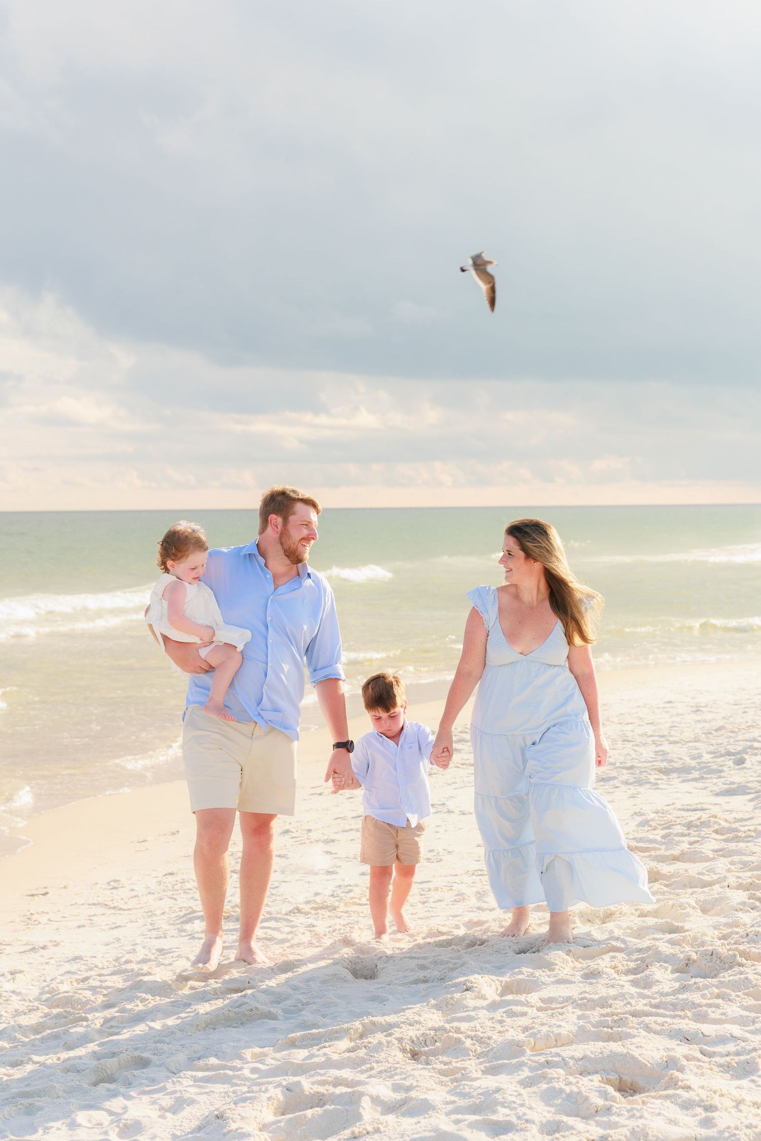 Happy family of four walking hand-in-hand on a sunny beach, with a seagull flying overhead in the cloudy sky.