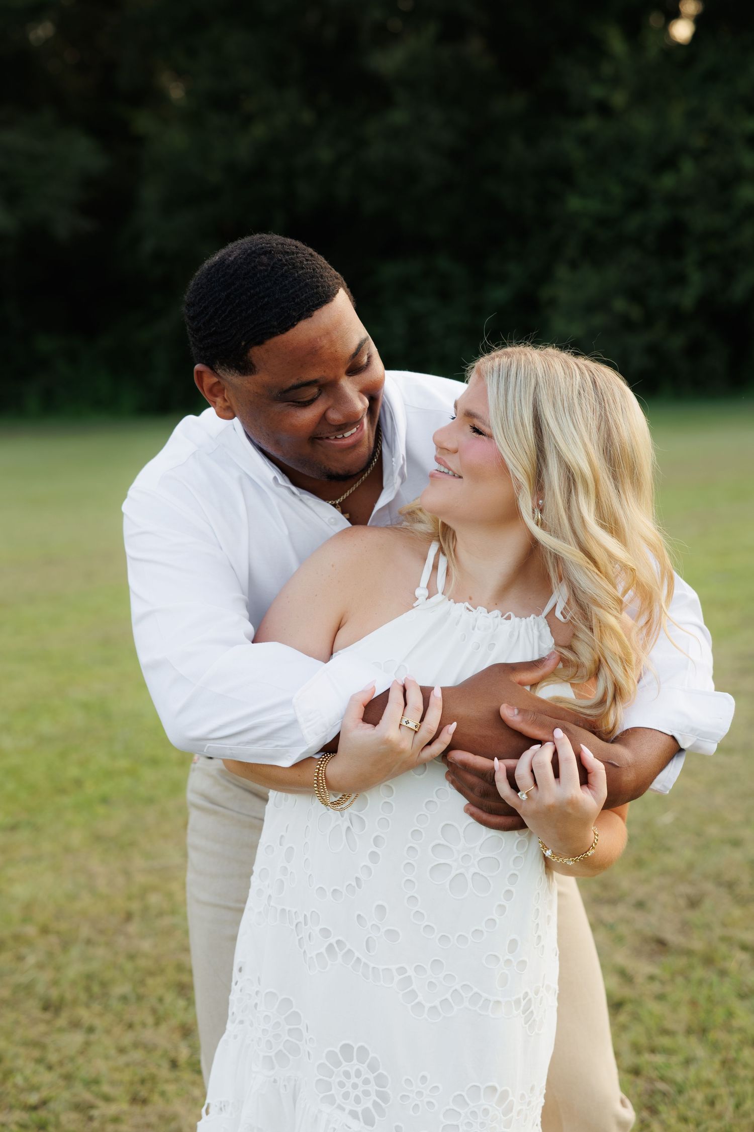 A smiling interracial couple embraces outdoors, the man in white shirt hugging a blonde woman in a white lace dress.