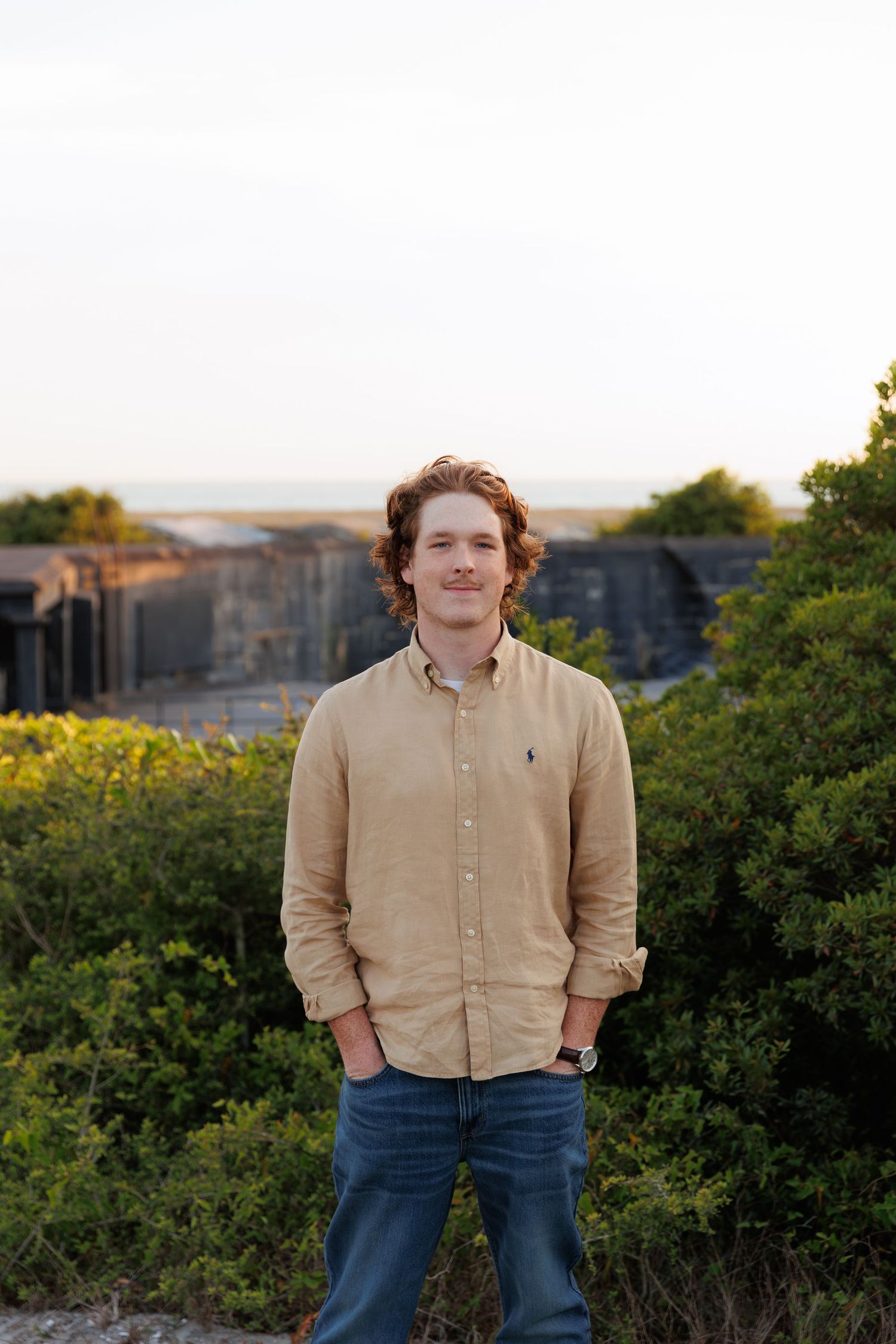 Young man in beige button-up shirt standing outdoors near historic fort ruins at dusk.