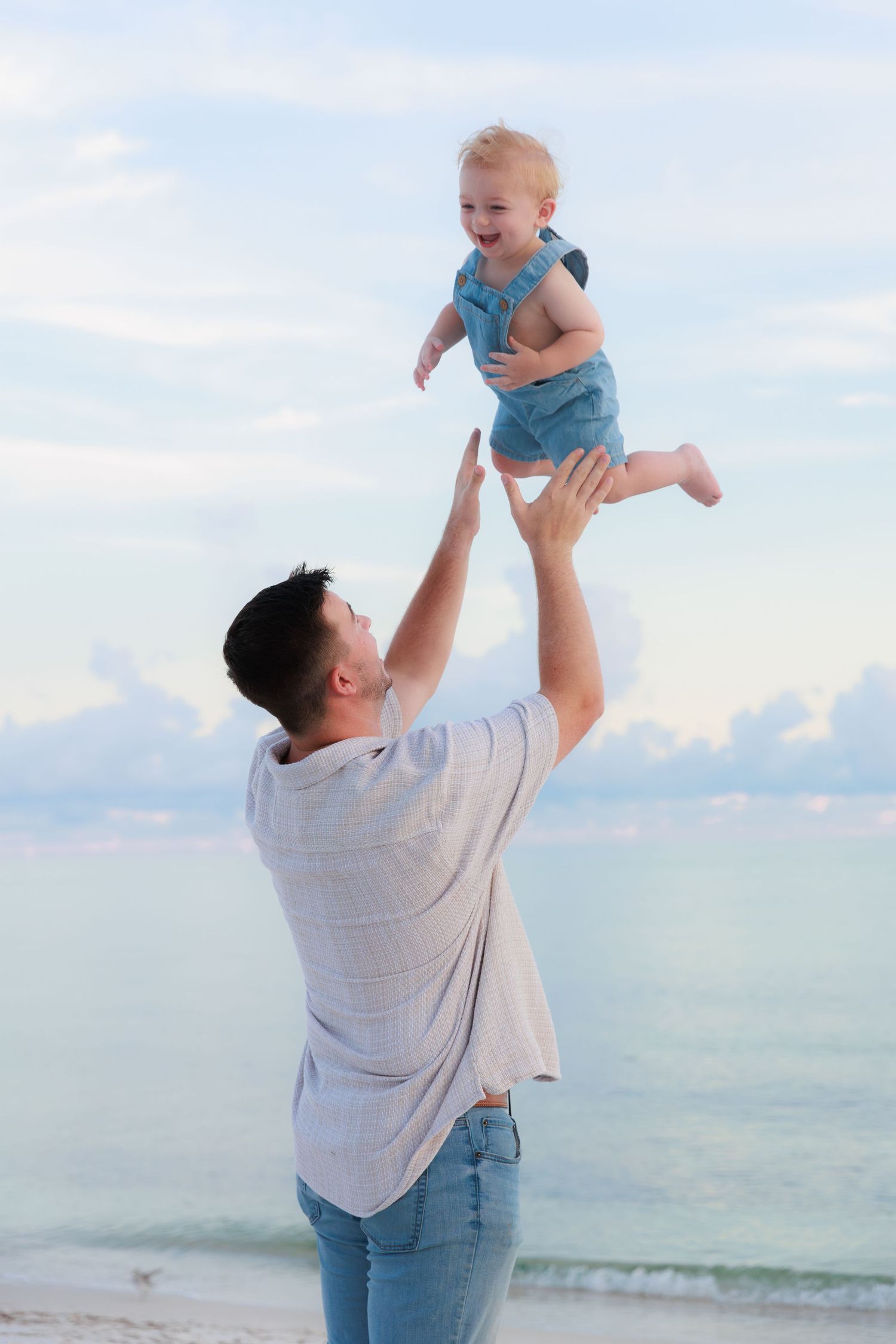Father joyfully lifting smiling baby in blue outfit high above his head on a serene beach at dusk.