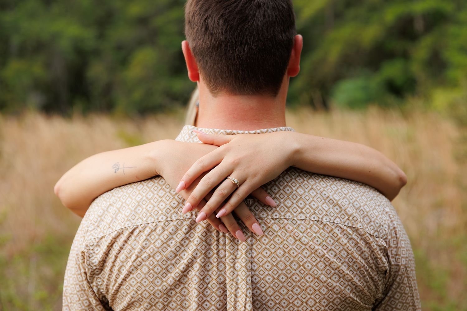 Couple embracing in golden field, woman's hand with ring on man's shoulder, lush green trees in background.