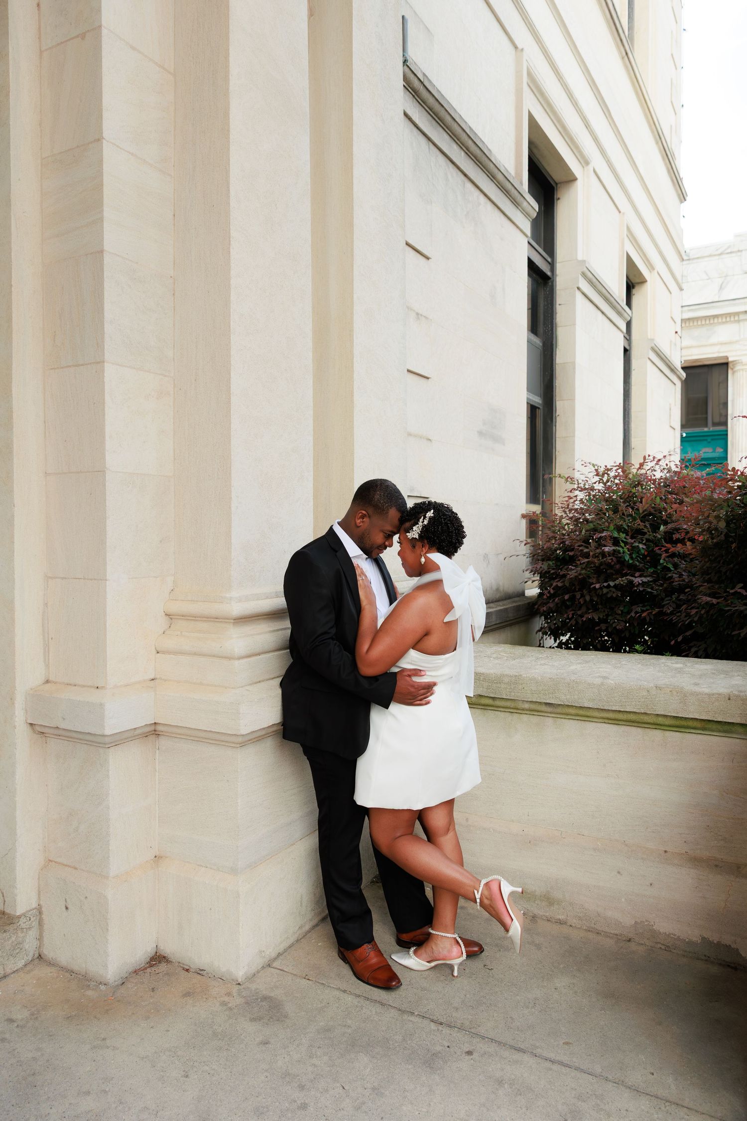 A Black couple shares a romantic embrace against a stone column, the bride in a short white dress and groom in a dark suit.