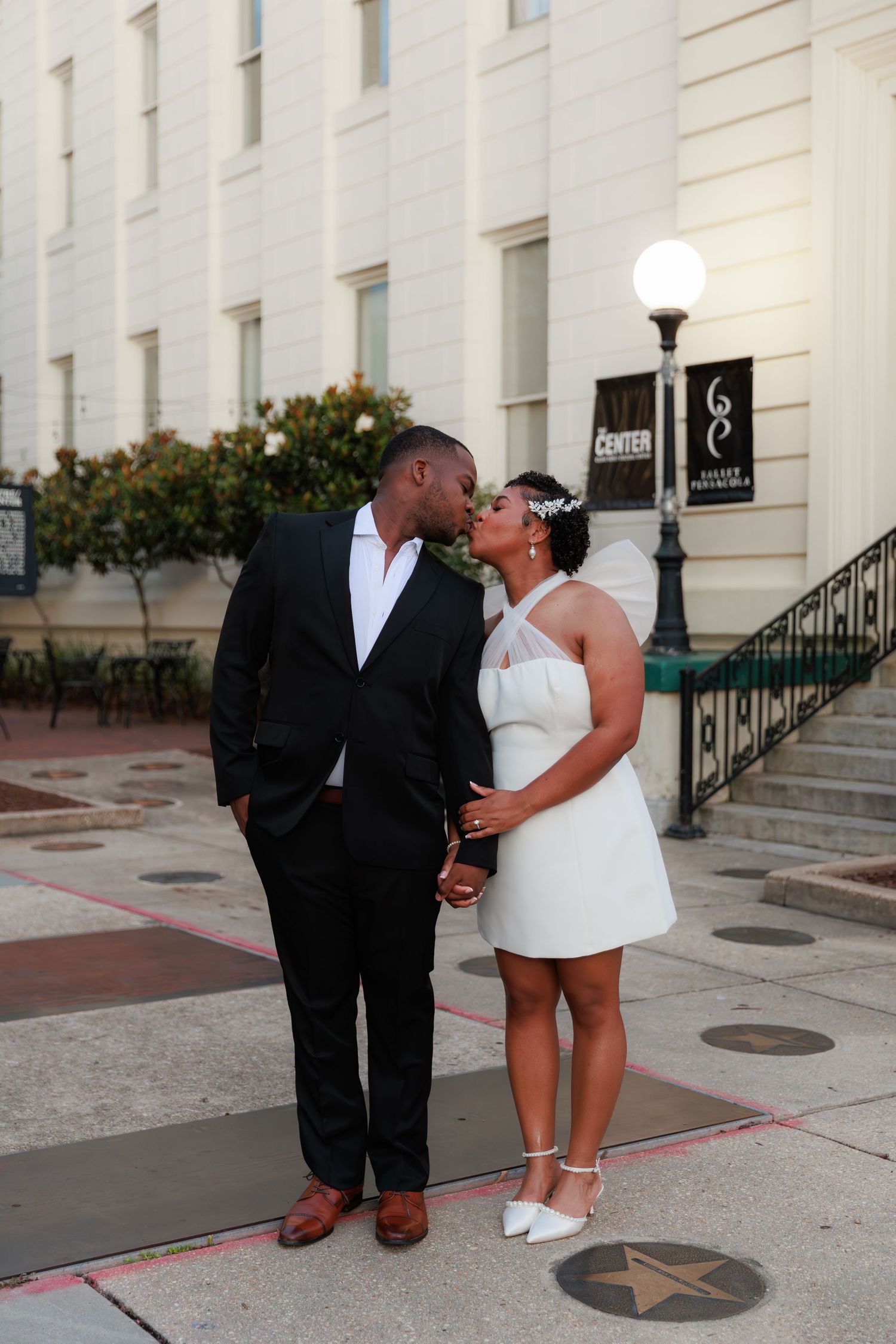 A couple shares a kiss outside a wedding venue, groom in black suit, bride in white mini dress and heels.