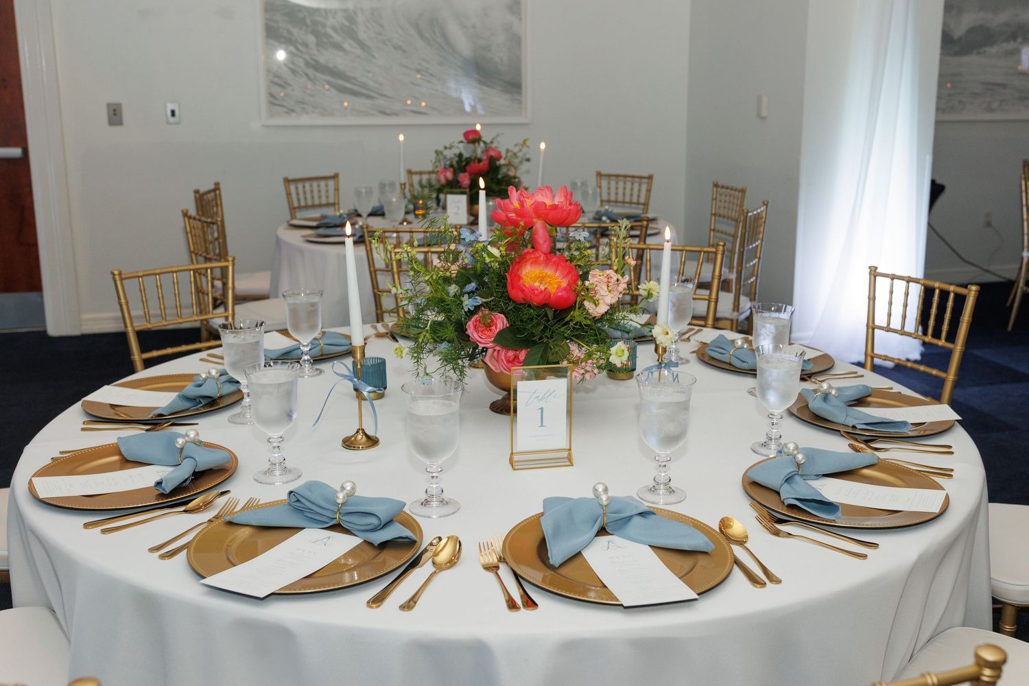 Elegant reception table settings with gold chargers, blue napkins, and white tablecloths at multiple rounds.